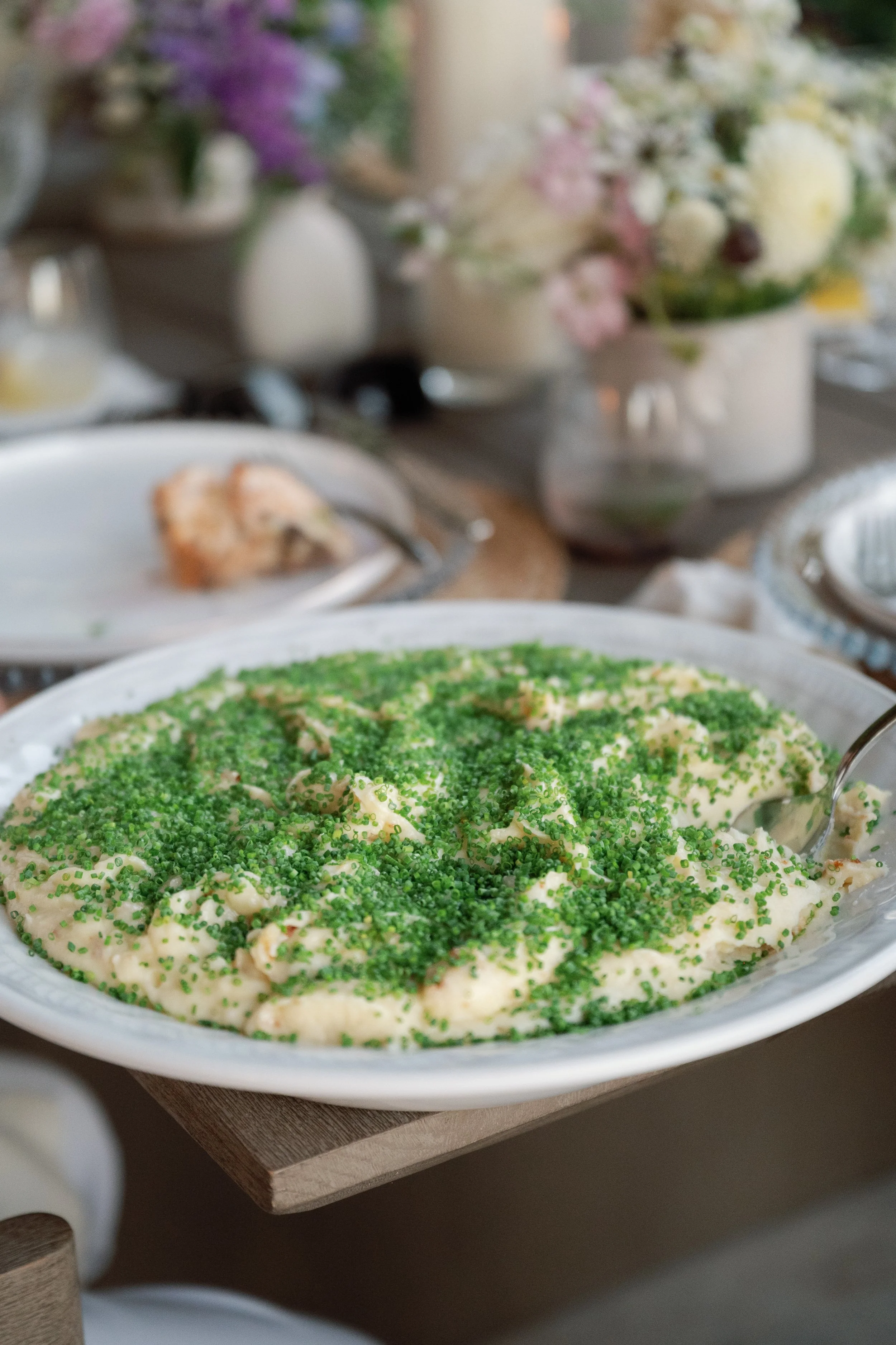 A dish covered with green sprinkles, likely food, on a white oval plate. In the background, there are blurred flowers and other tableware.