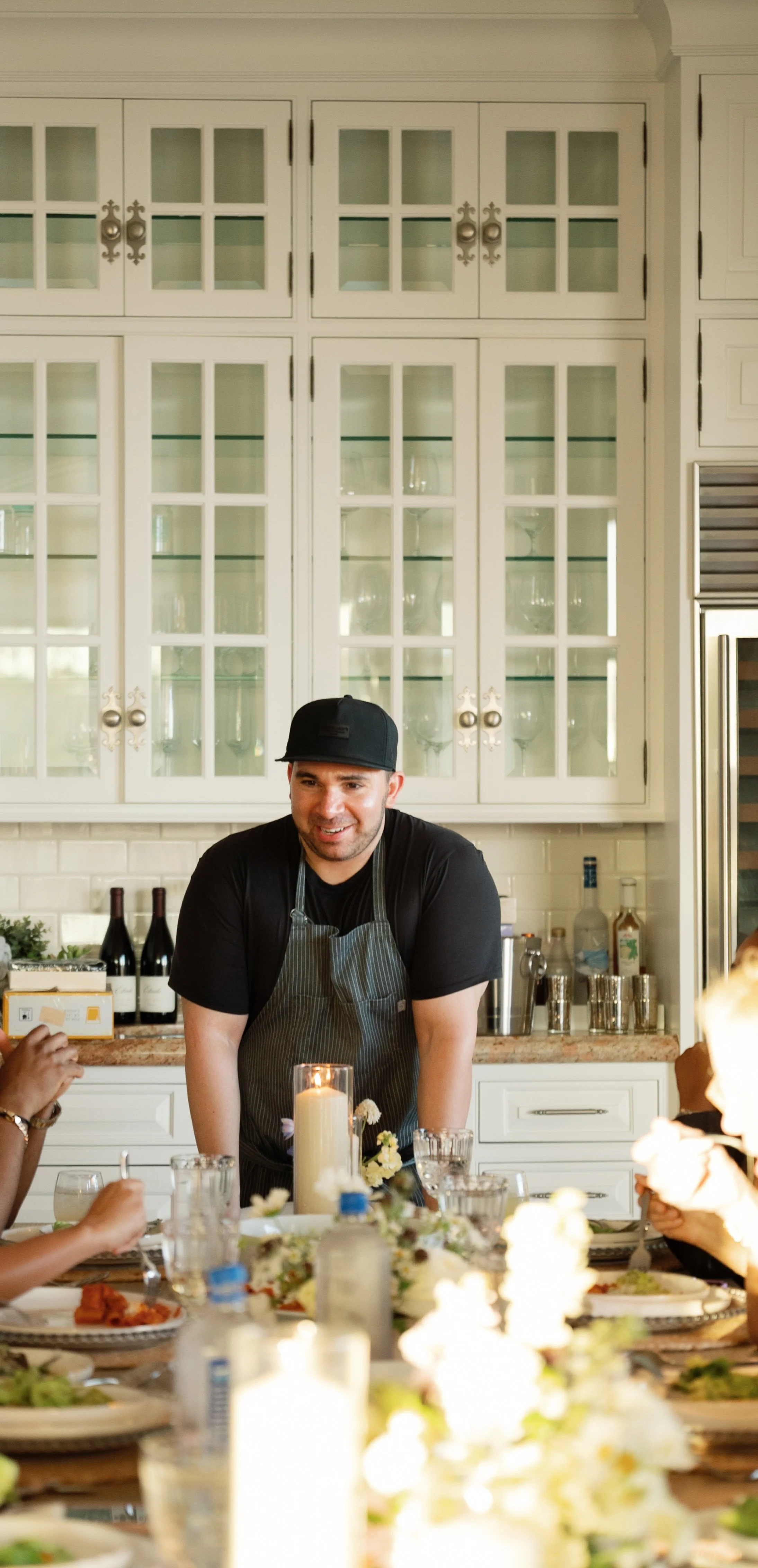 A smiling man wearing a black hat and apron stands at a dining table with dishes, glasses, and a lit candle, in a white kitchen with glass-front cabinets in the background.