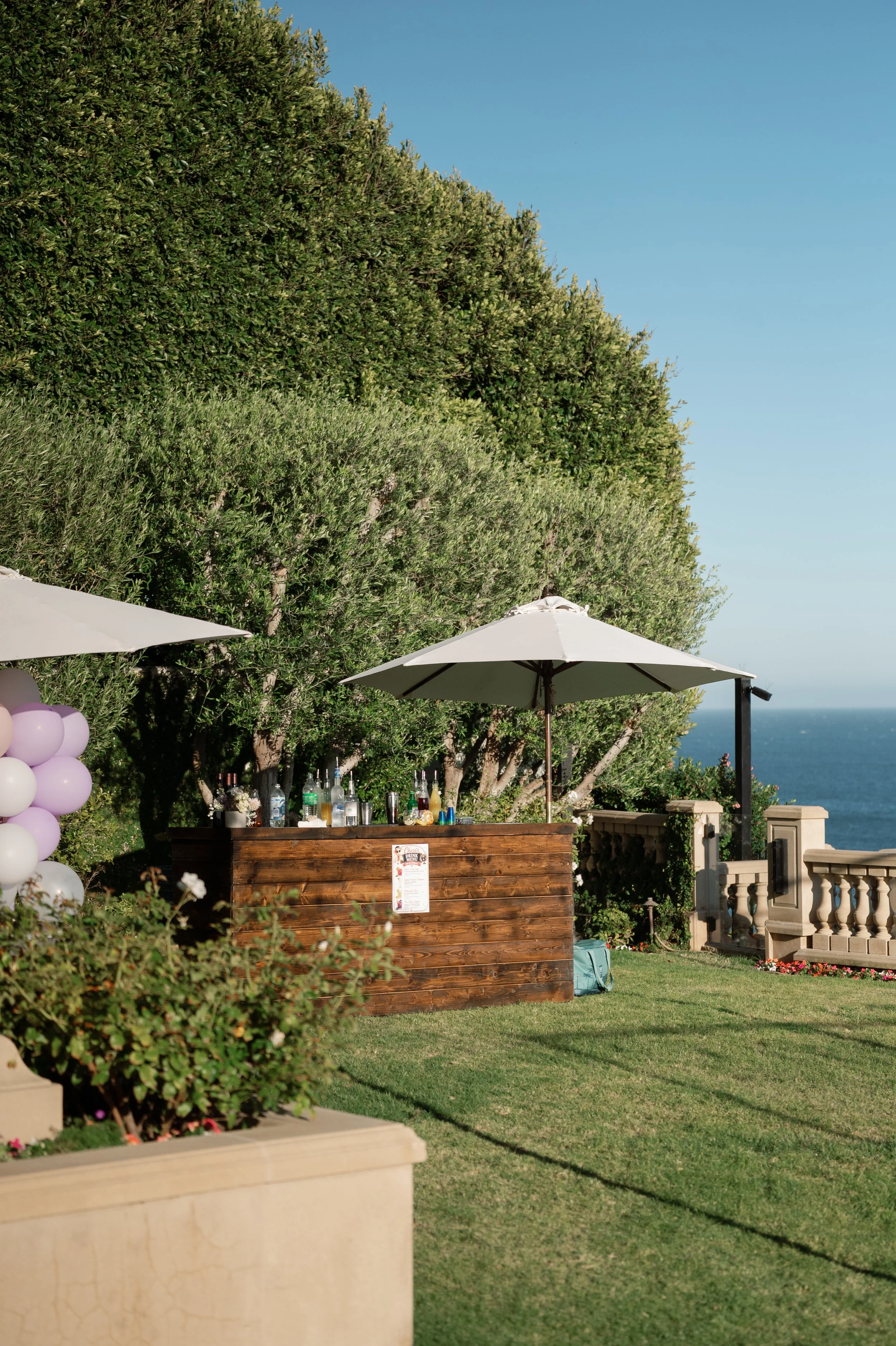 Outdoor bar setup with a wooden counter, surrounded by trees and greenery, under large patio umbrellas, overlooking the ocean in the distance.