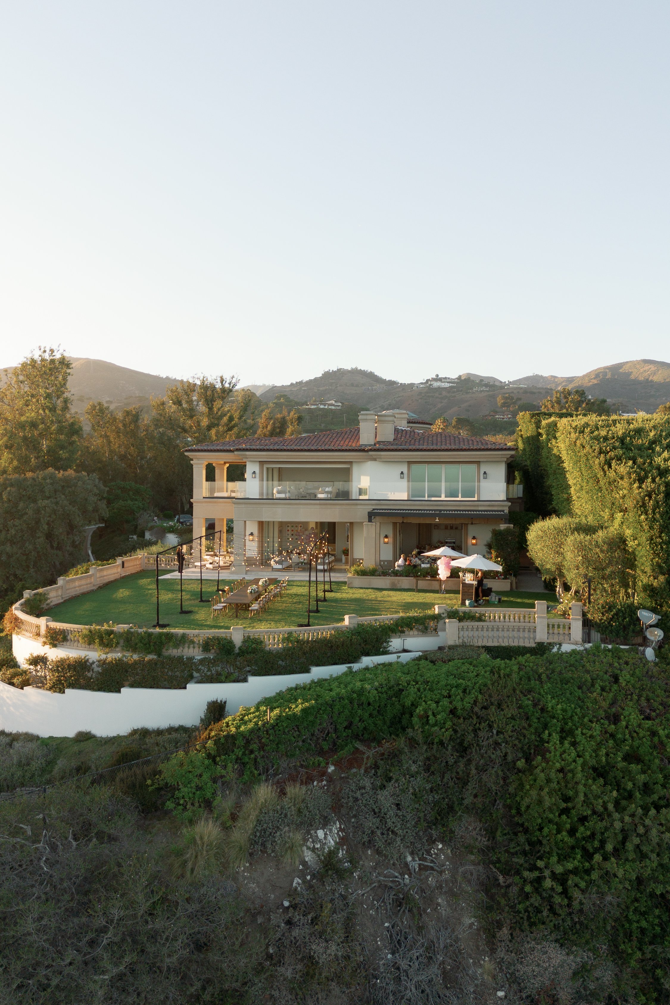 A modern house with a balcony, surrounded by trees and bushes, with mountains in the background under a clear sky.