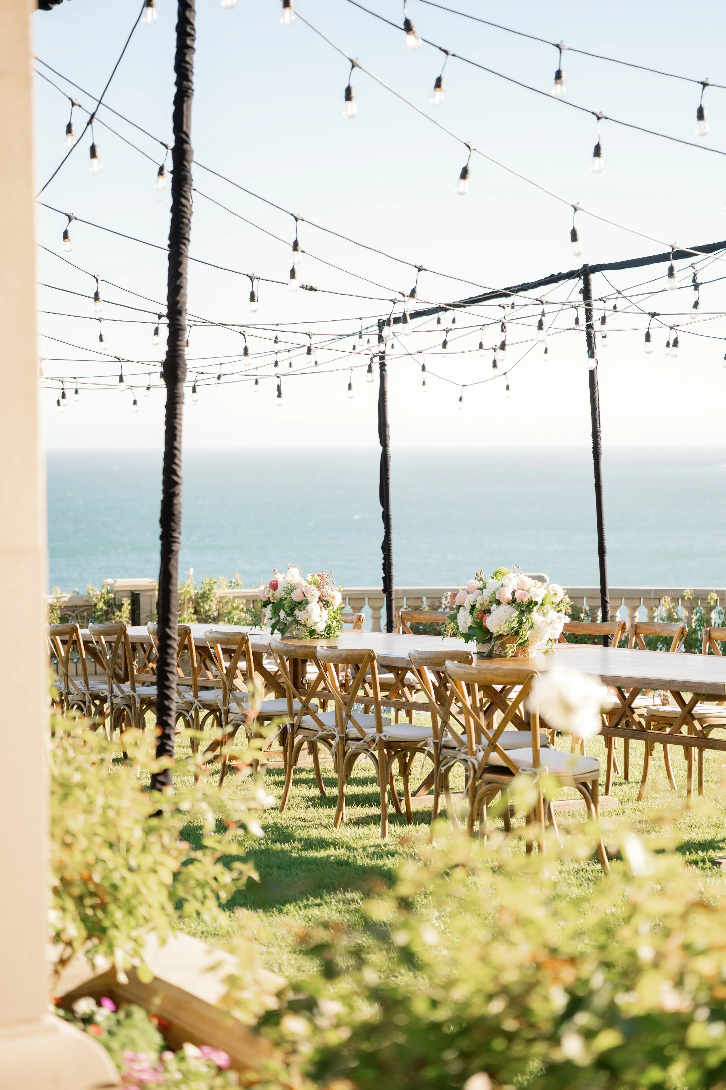 Outdoor event setup with long wooden tables, floral centerpieces, string lights overhead, ocean view in the background, and green grass in the foreground.