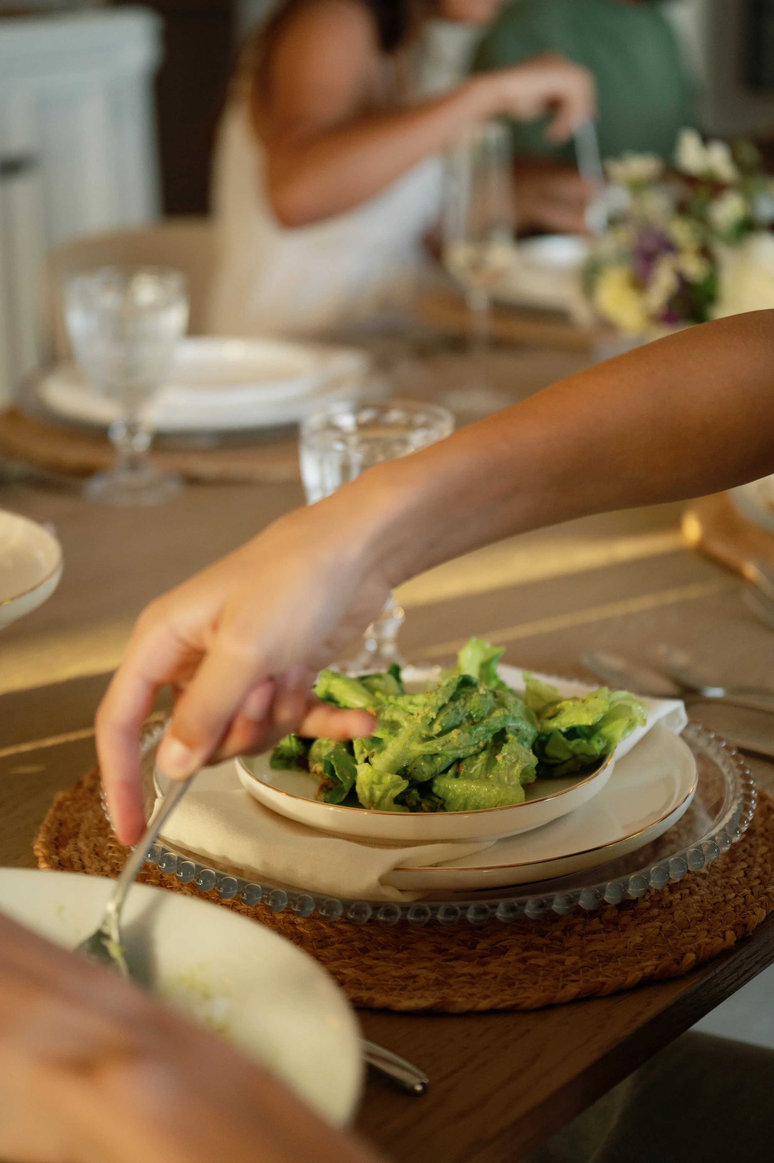 Person serving salad onto a plate at a dining table with water glasses and a floral centerpiece in the background.