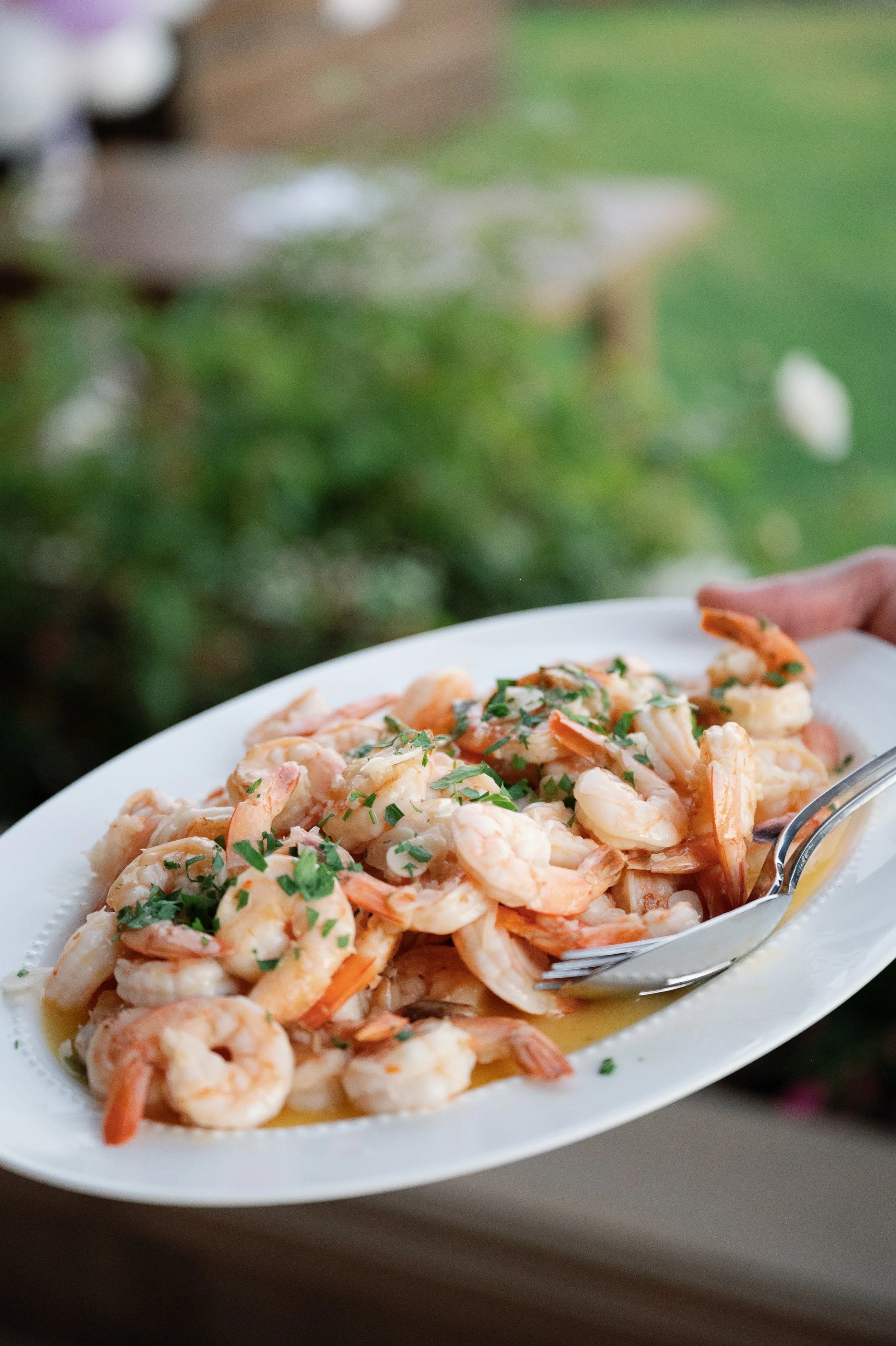 A white oval-shaped plate with cooked shrimp garnished with chopped herbs, possibly parsley, and a lemon wedge on the side. The background is blurred with green and brown colors, suggesting an outdoor setting.