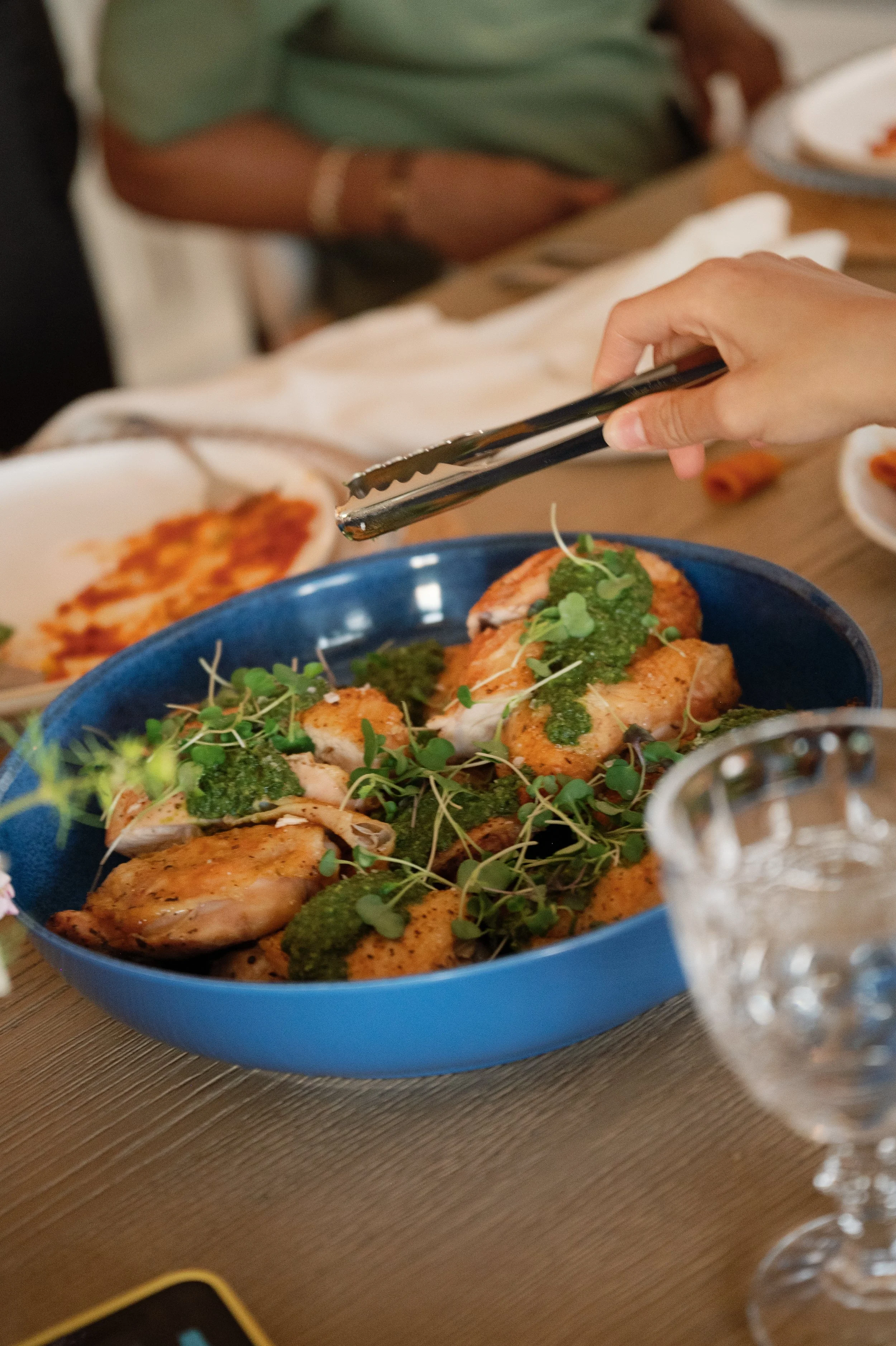 A blue bowl of roasted chicken topped with green herbs and microgreens on a wooden table, with a person's hand holding tongs, and plates with other food in the background.