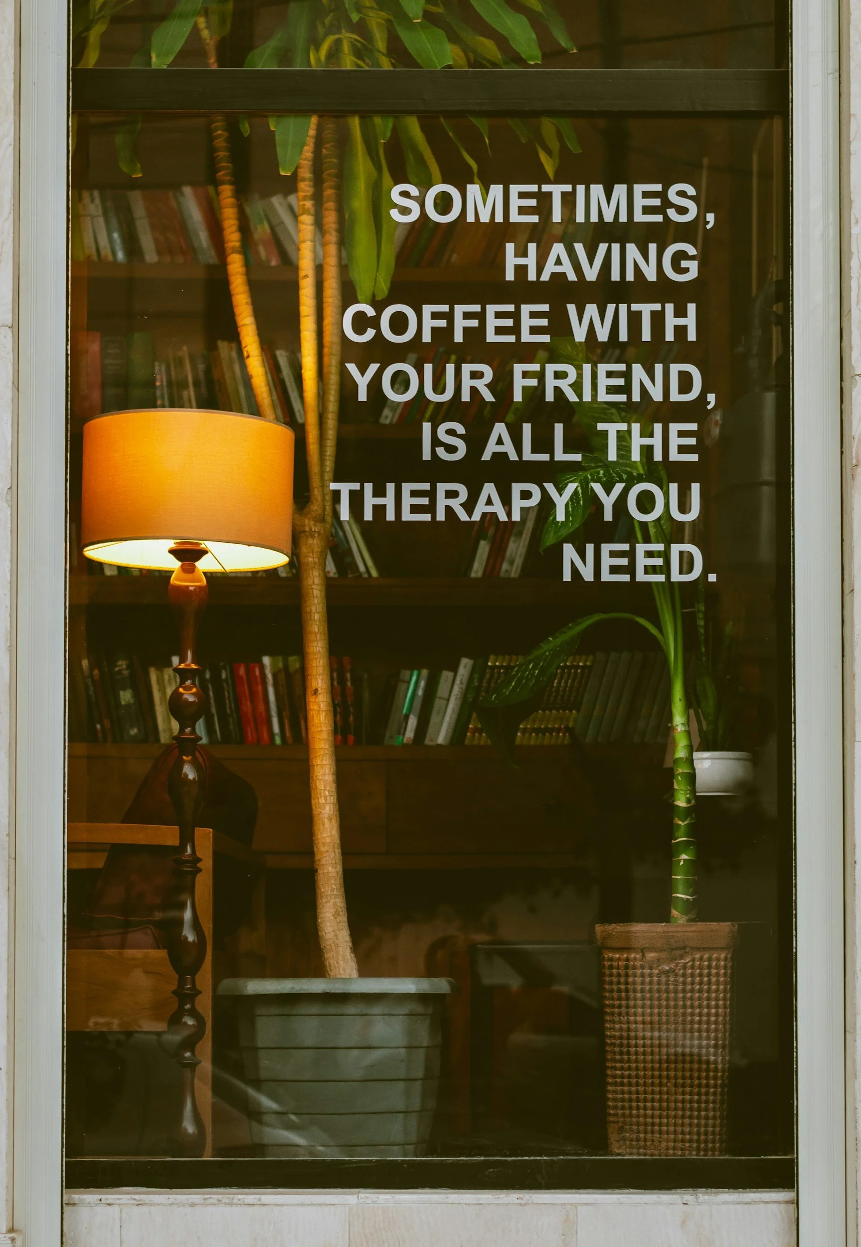 Glass pane featuring text "sometimes coffee with a friend is all the therapy you need" in a cozy, warm, office with books.