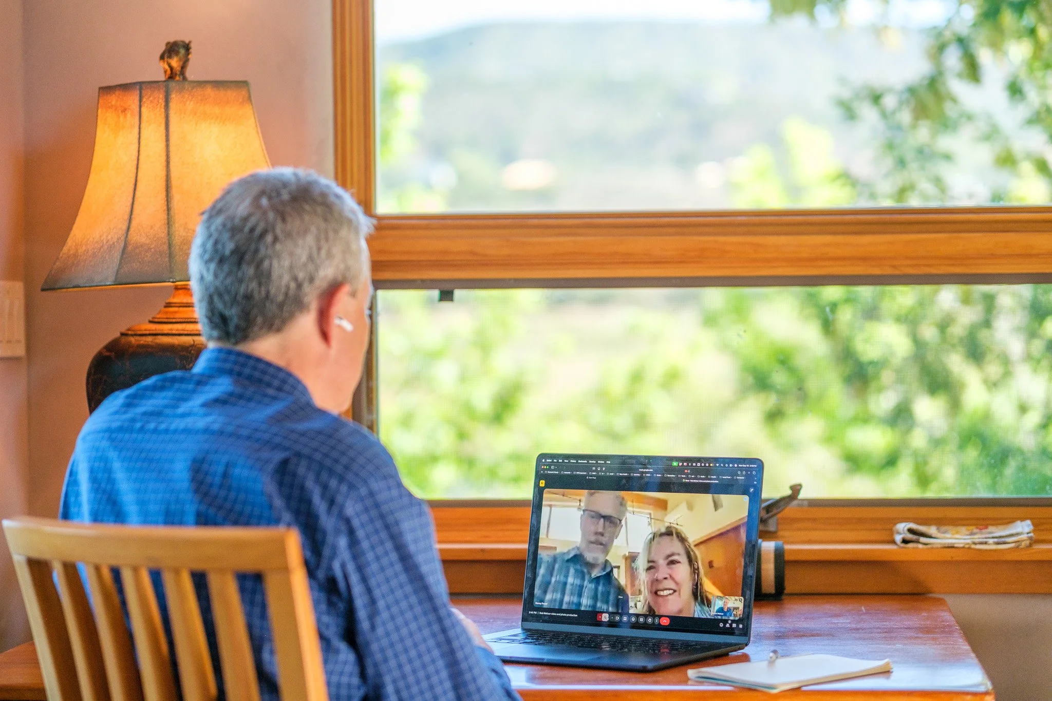Man with gray hair sitting at a wooden desk, participating in a video call on a laptop. The laptop screen shows two smiling people, a man and a woman, chatting. The room has a large window with green trees outside and a lamp on the left side.