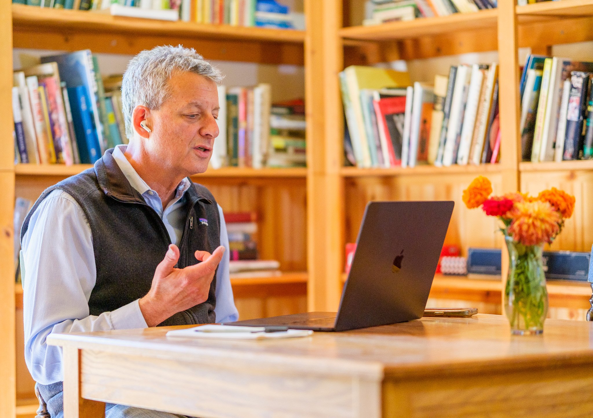 A middle-aged man with gray hair, wearing a blue shirt and black vest, sitting at a wooden table in a room with wooden bookshelves filled with books. He is engaged in a video call, wearing wireless earbuds, and gesturing with his hand. There is a laptop, a notebook, and a vase of orange and pink flowers on the table.