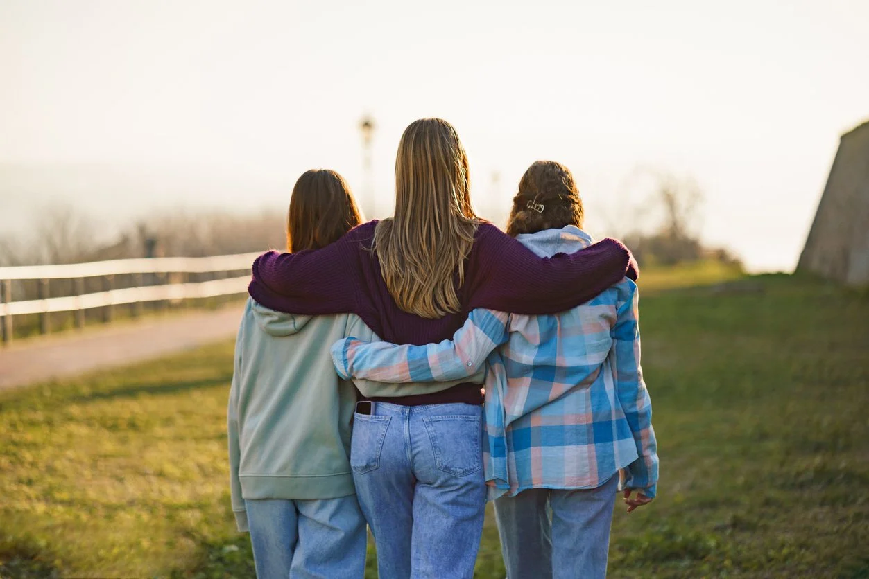 Three young women walking together outside, with their arms around each other, during sunset.