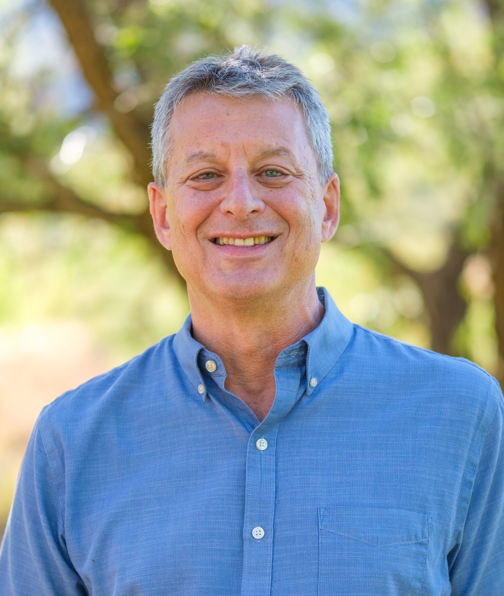 A smiling middle-aged man with gray hair wearing a blue button-up shirt outdoors with trees and greenery in the background.