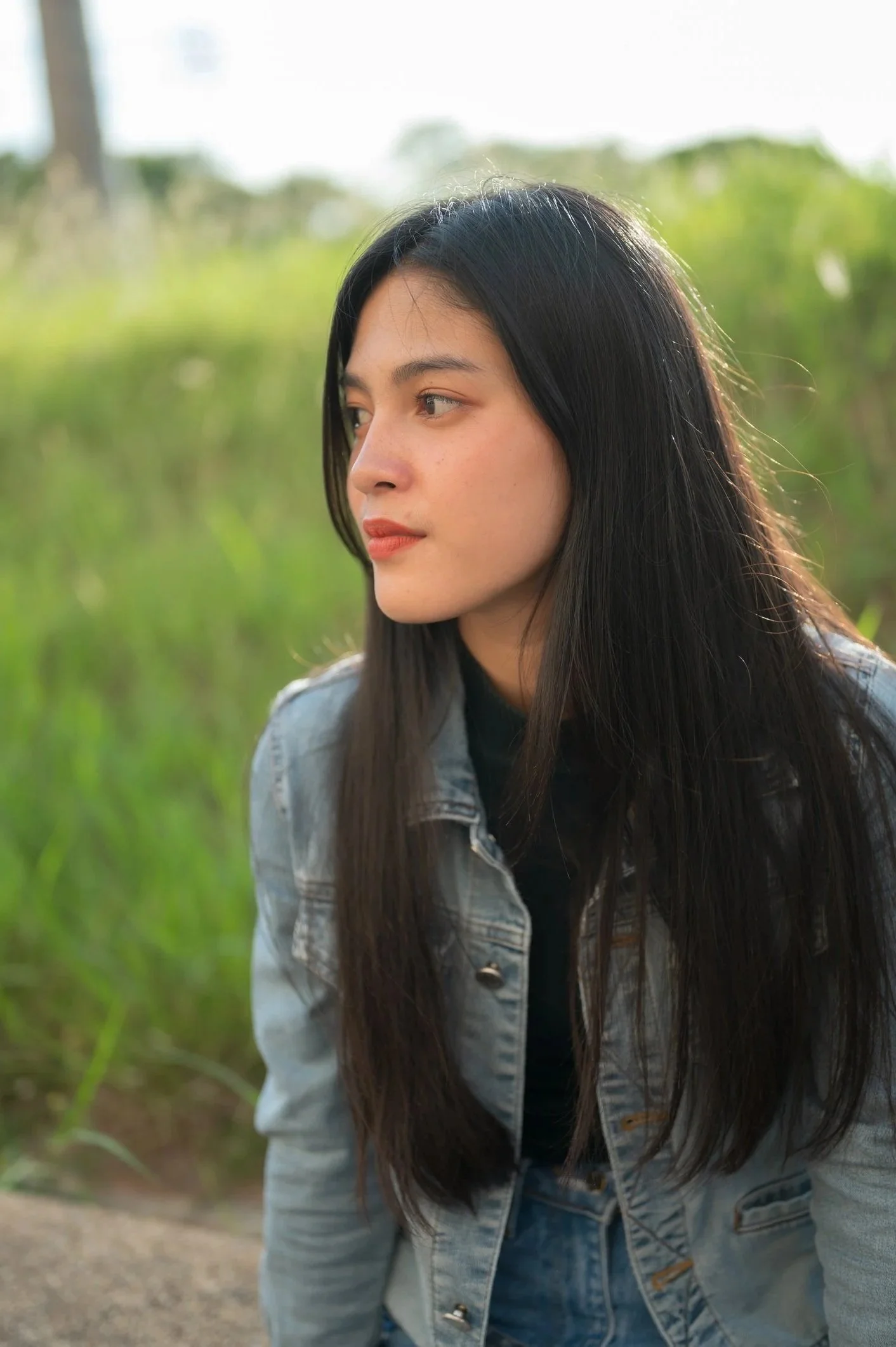 A young woman with long dark hair and light skin, wearing a denim jacket, looking to the left outdoors with blurred greenery in the background.
