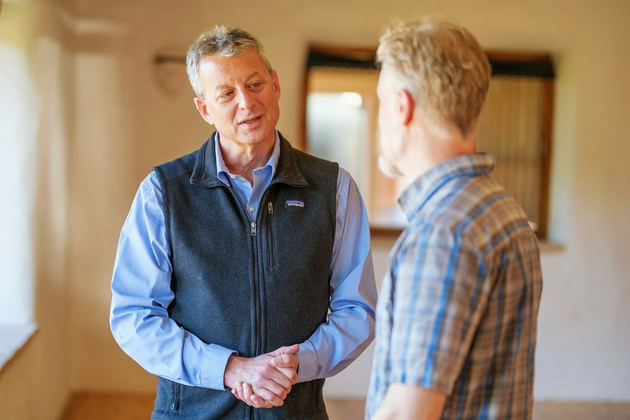 Two men talking indoors, one in a blue shirt and black vest, speaking to another man in a plaid shirt.