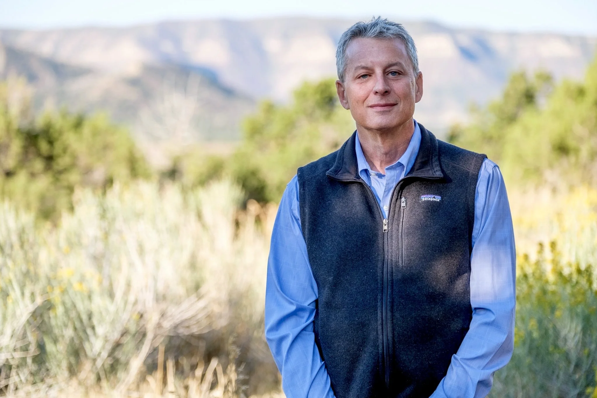 A middle-aged man with gray hair outdoors, wearing a blue shirt and a black Patagonia vest, standing in front of green foliage and mountains in the distance.