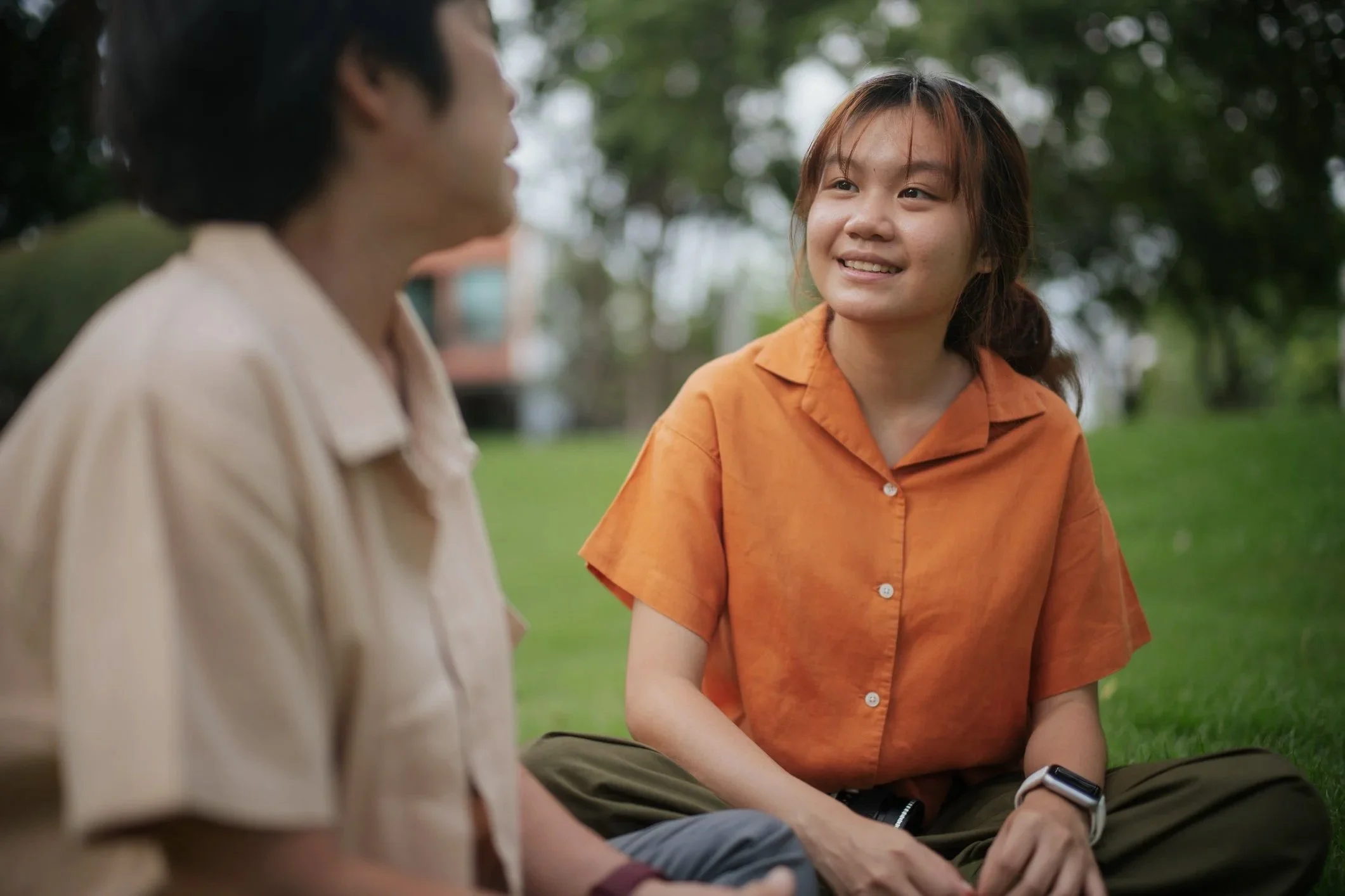 Two women sitting on grass in a park, engaging in conversation, with trees and a building in the background.