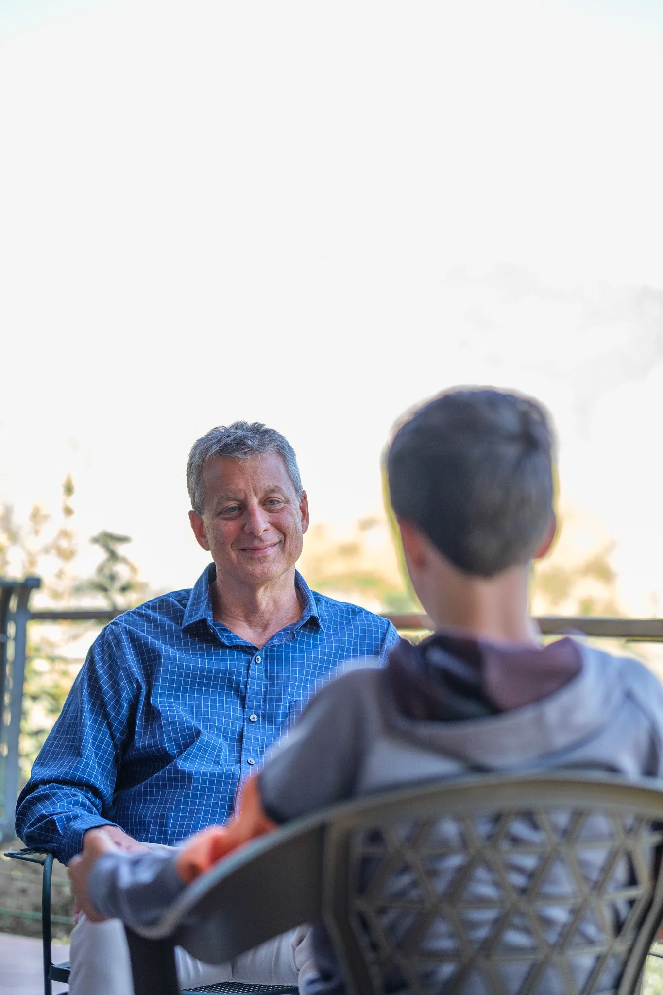 An older man and a boy are sitting outdoors, facing each other; the man is smiling at the boy who has his back to the camera.