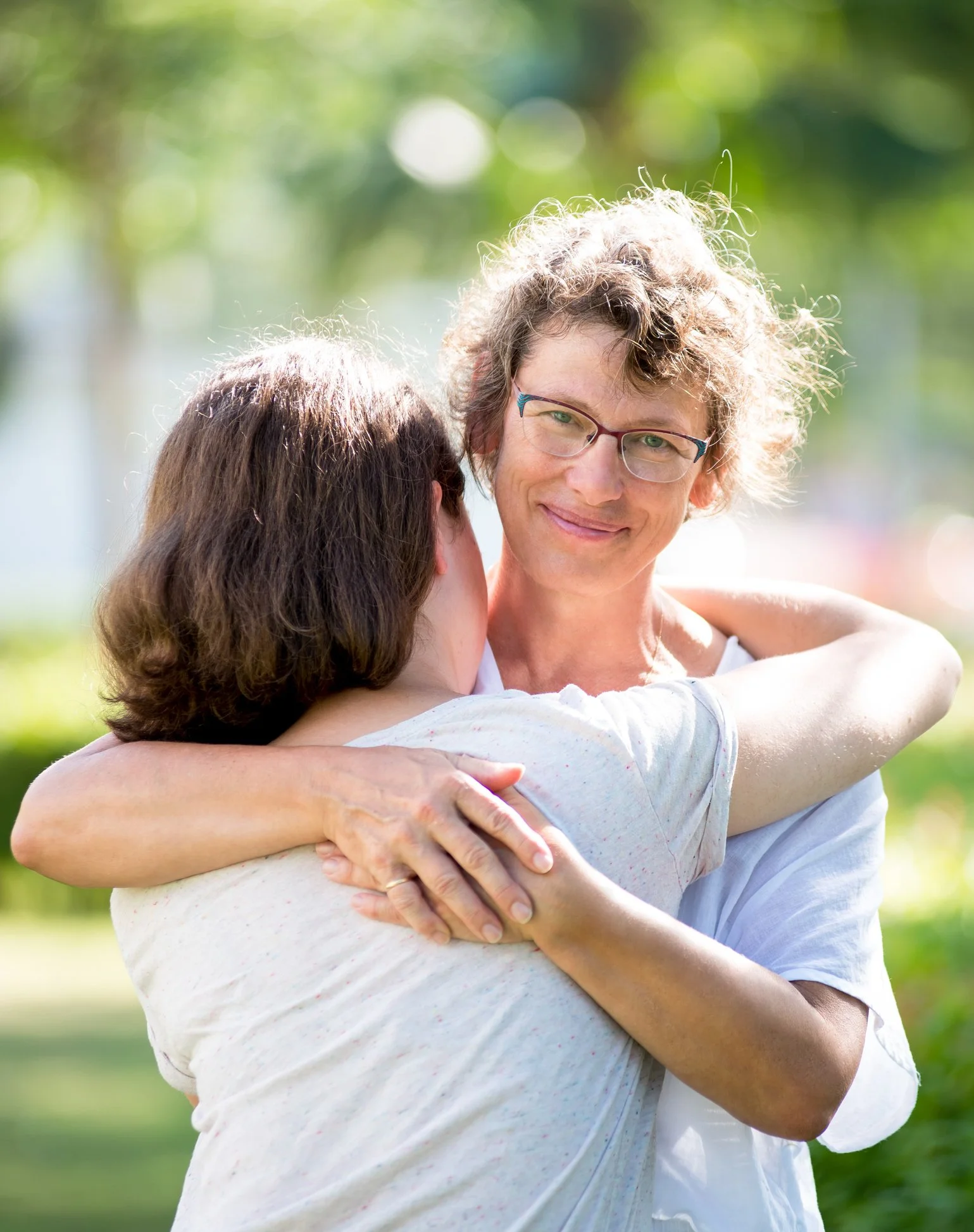 A man with curly hair and glasses hugging a woman outdoors on a sunny day, with a blurred green background.