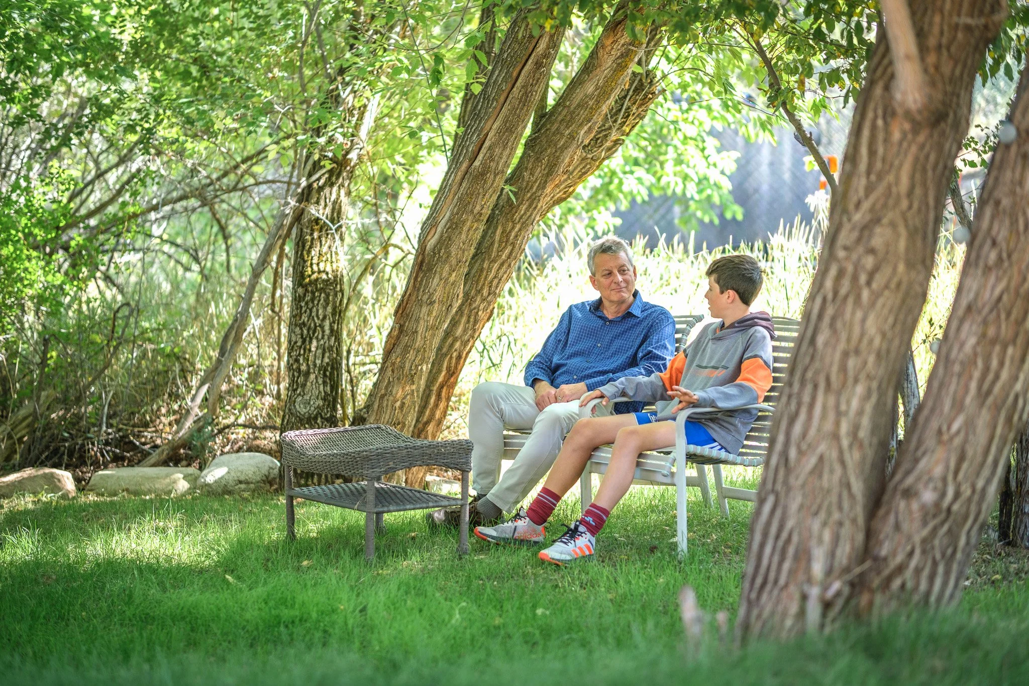 An older man and a young boy sitting on a bench in a shaded backyard with trees and grass, engaged in conversation.