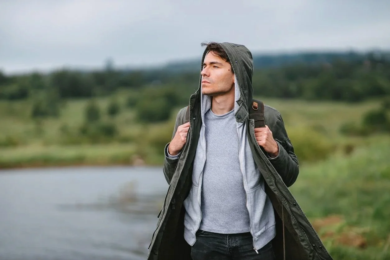 Man in gray sweater and black jacket with hood and backpack standing outdoors near a body of water, looking into the distance.