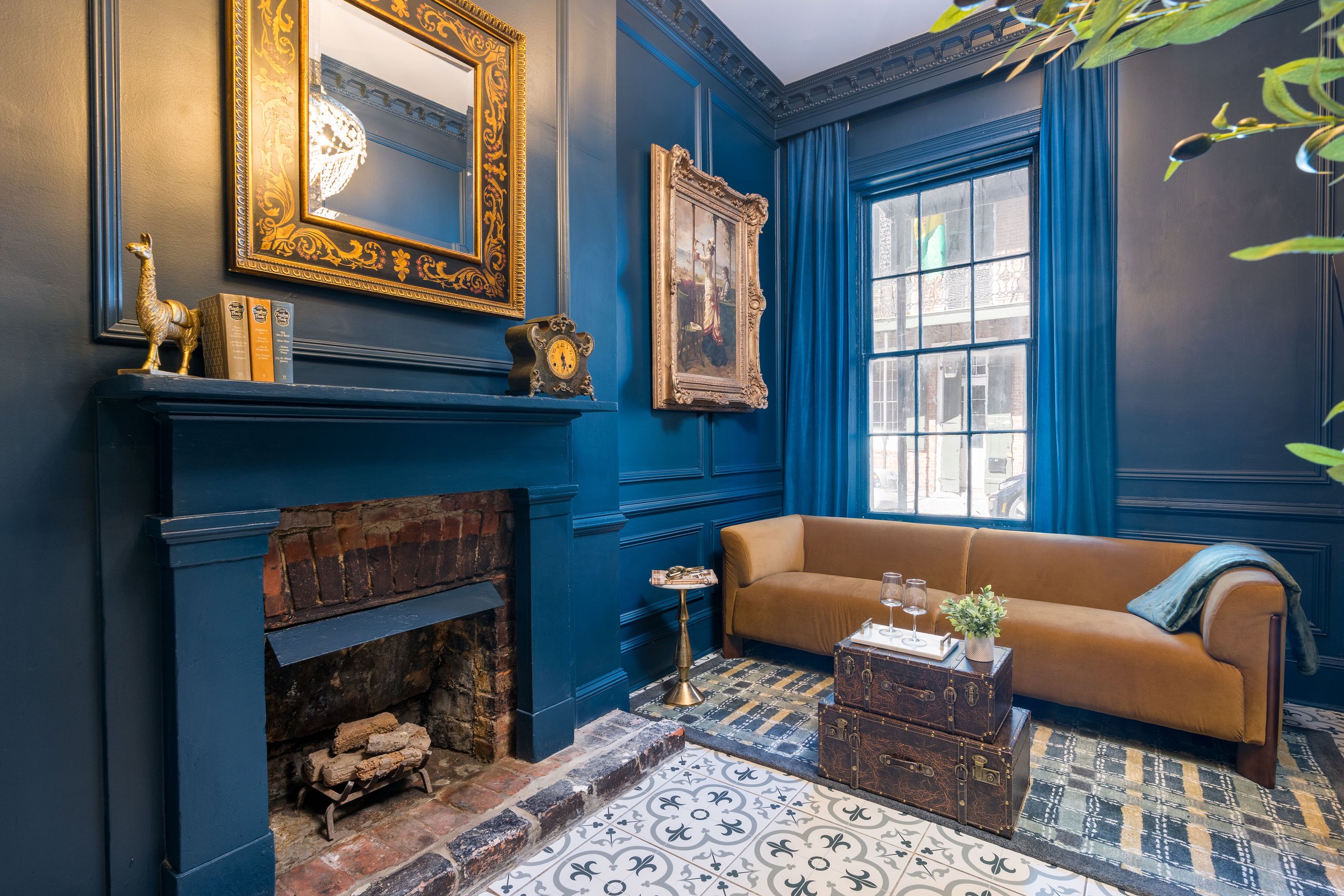 A cozy living room with dark blue walls, a mustard yellow sofa, a patterned rug, and large window with blue curtains. There is a brick fireplace, a decorative mirror above it, and framed artwork on the walls. A small side table holds glasses and a potted plant, and vintage suitcases serve as a coffee table.