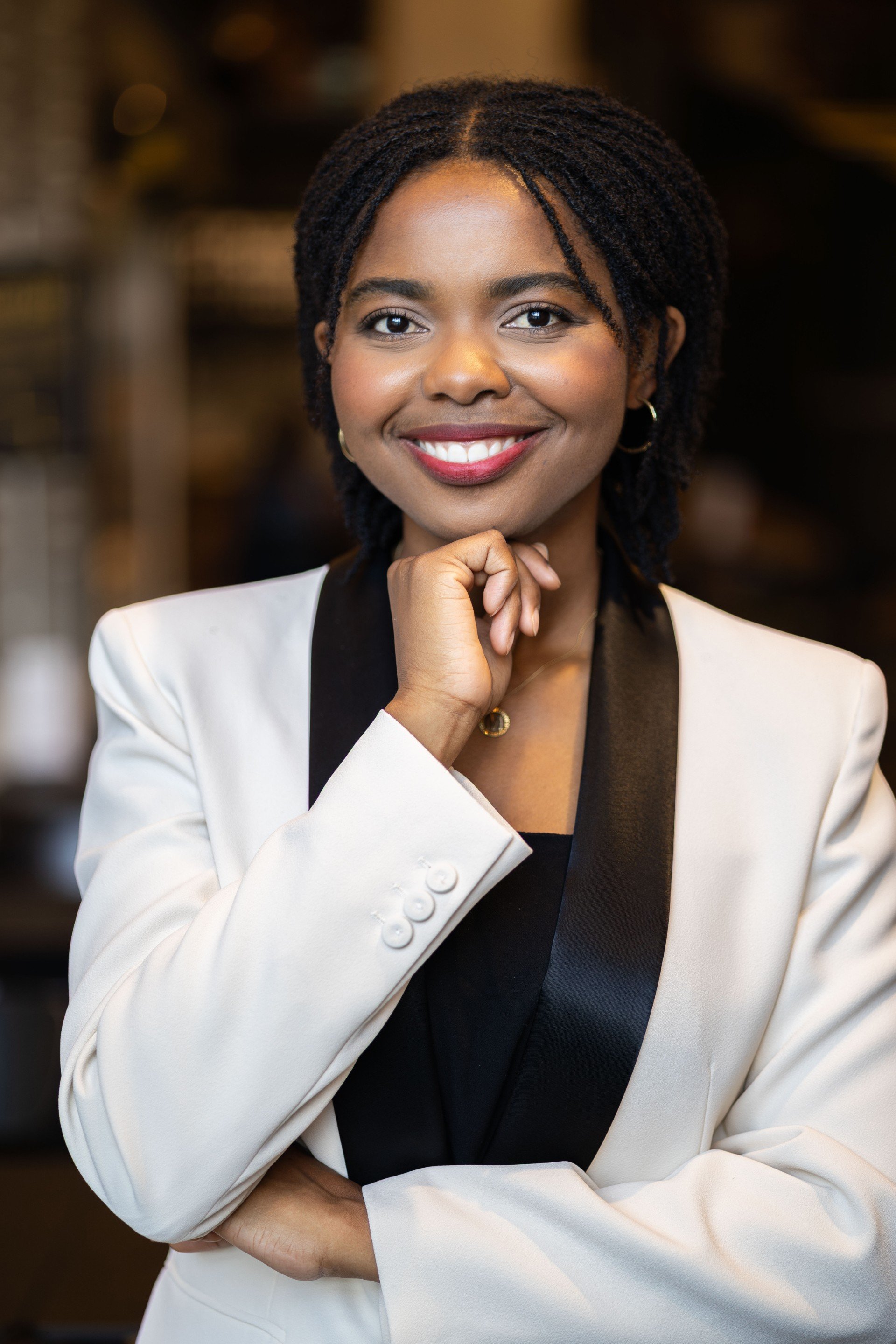 Maphrida Wehrli - A woman with medium dark skin tone and styled hair, wearing a cream blazer with black lapel and a black top, smiling and resting her chin on her hand in an indoor setting.