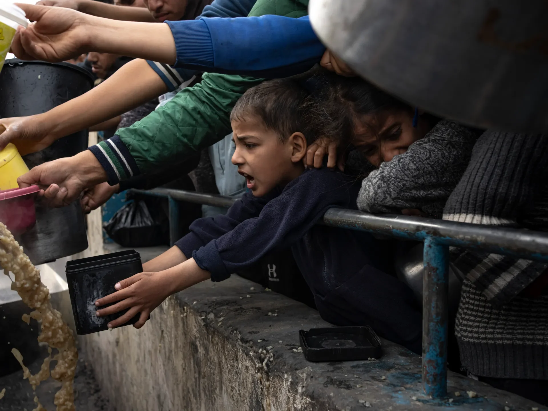 A group of people, including children, are reaching over a metal railing to collect food or water from a container held by someone. The children look distressed and are stretching to grab the contents, with some showing expressions of urgency and discomfort.
