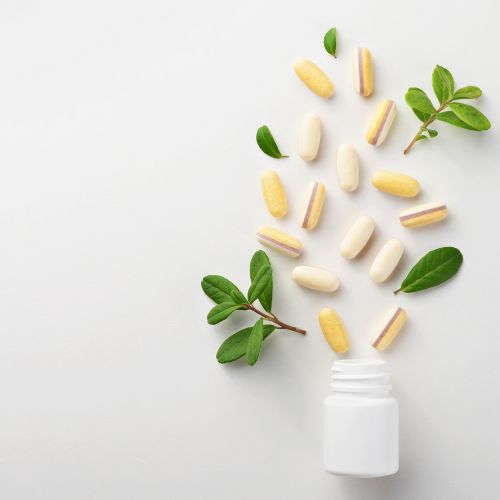 Various colorful chewable vitamins and green leaves spilling from a white pill bottle on a white surface.