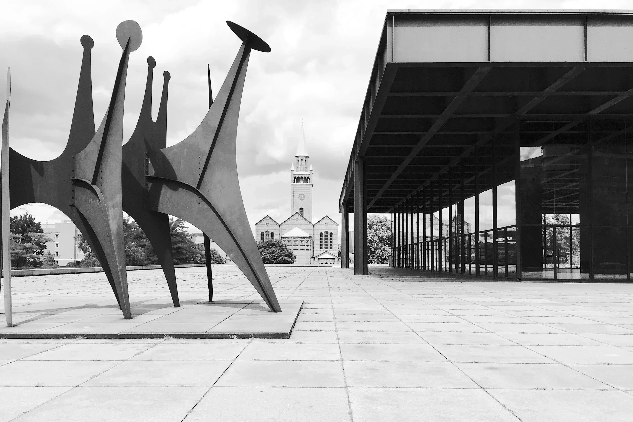 Moderne Skulptur auf einem freien Platz mit einem historischen Kirchturm im Hintergrund, überdachte Glasfassade auf der rechten Seite, schwarz-weiß Aufnahme.