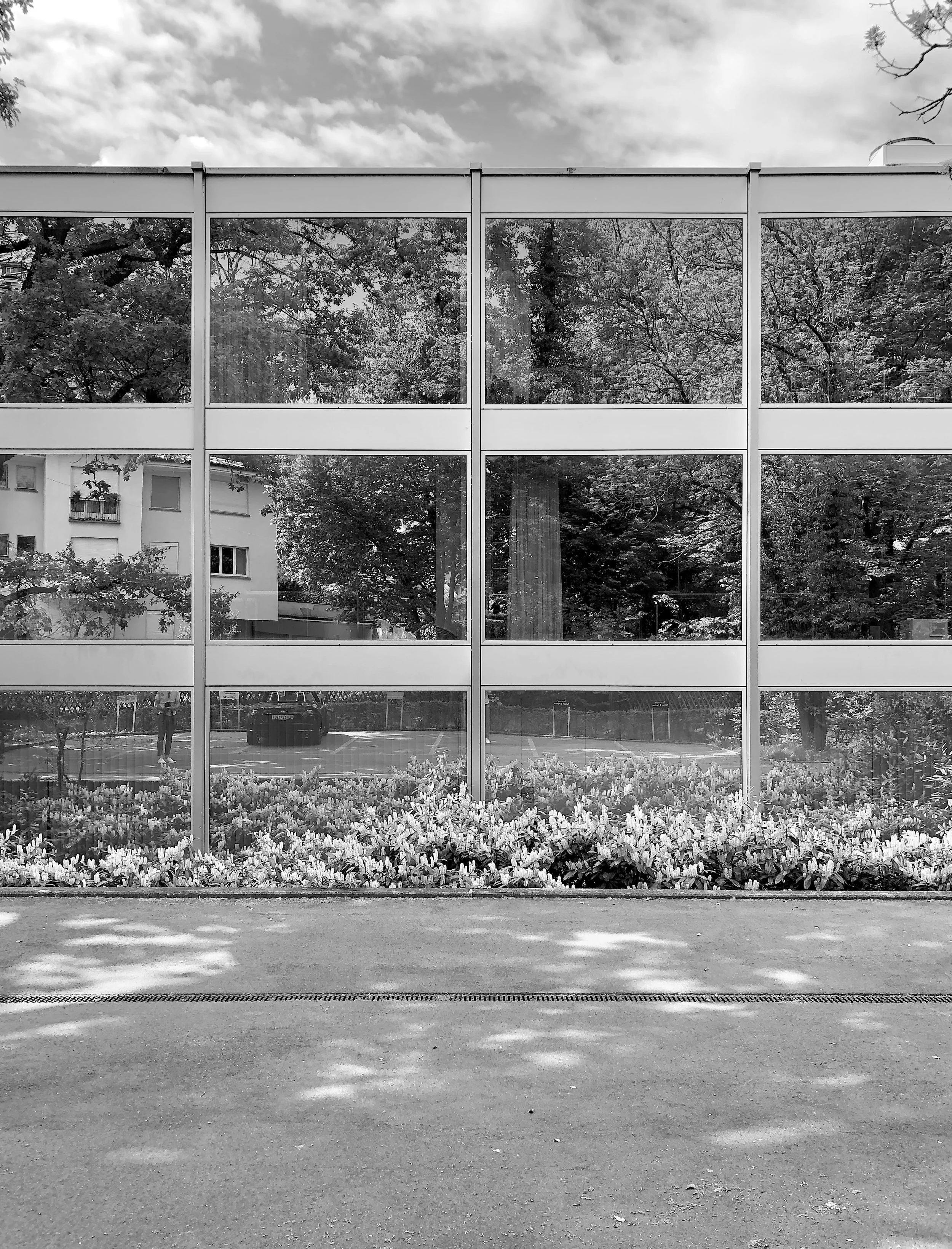 Ein modernes Gebäude mit großen Glasfenstern, dahinter Bäume und ein Himmel mit Wolken, im Vordergrund ein gepflasterter Bereich mit Blumenrabatten.