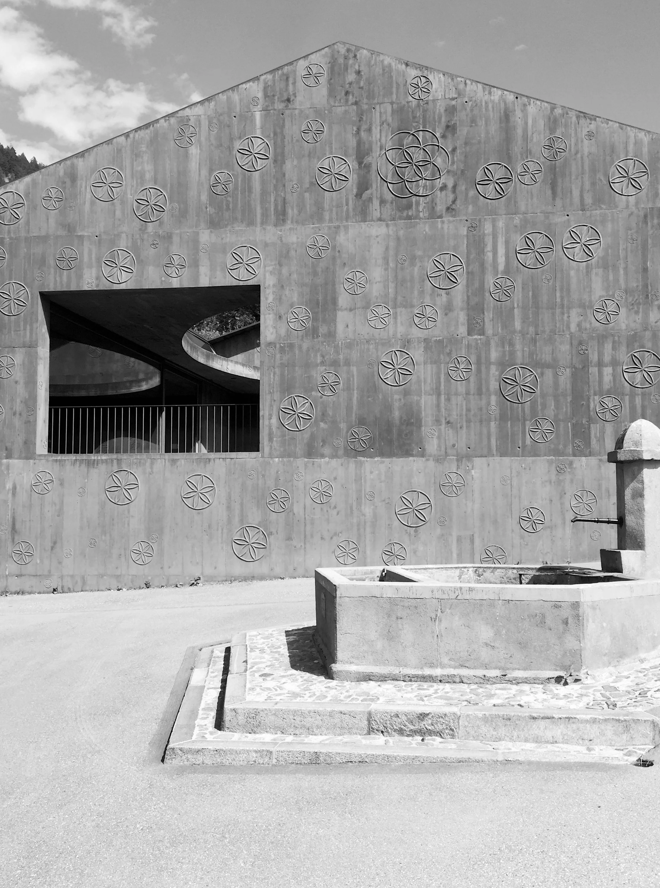 Ein modernes graues Betongebäude mit einem rechteckigen Loch im oberen Bereich und dekorativen Kreisen an der Fassade. Im Vordergrund ein Brunnen aus Stein.