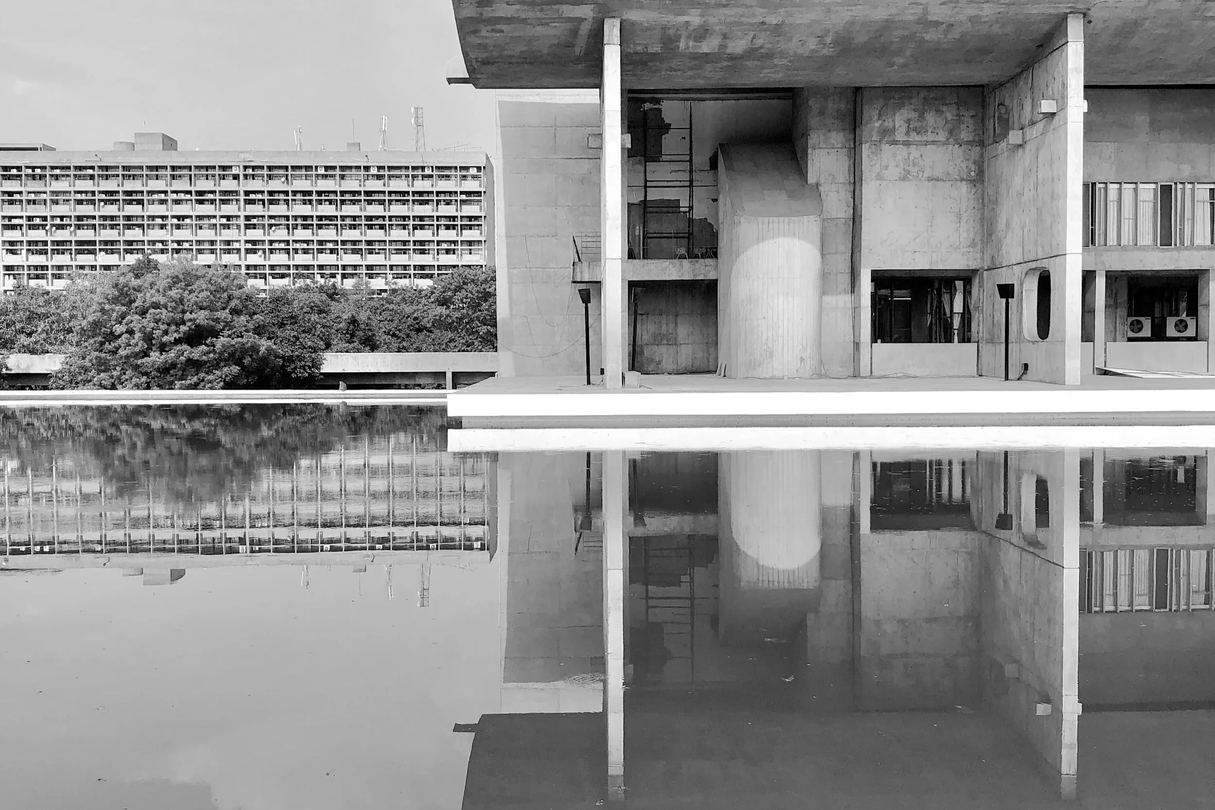 Ein modernes, skulpturales Gebäude aus Beton mit großen, runden und rechteckigen Öffnungen, neben einer Wasserfläche, die die Struktur spiegelt, und einem Hintergrund mit einem Wohnblock und Bäumen.