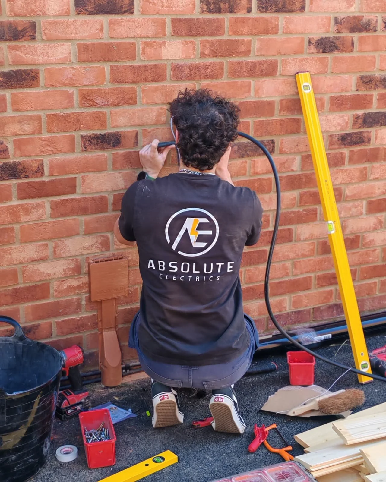 A person working on electrical wiring outside a brick wall, kneeling on the ground with tools and equipment around them, wearing a shirt with the logo 'Absolute Electric'.