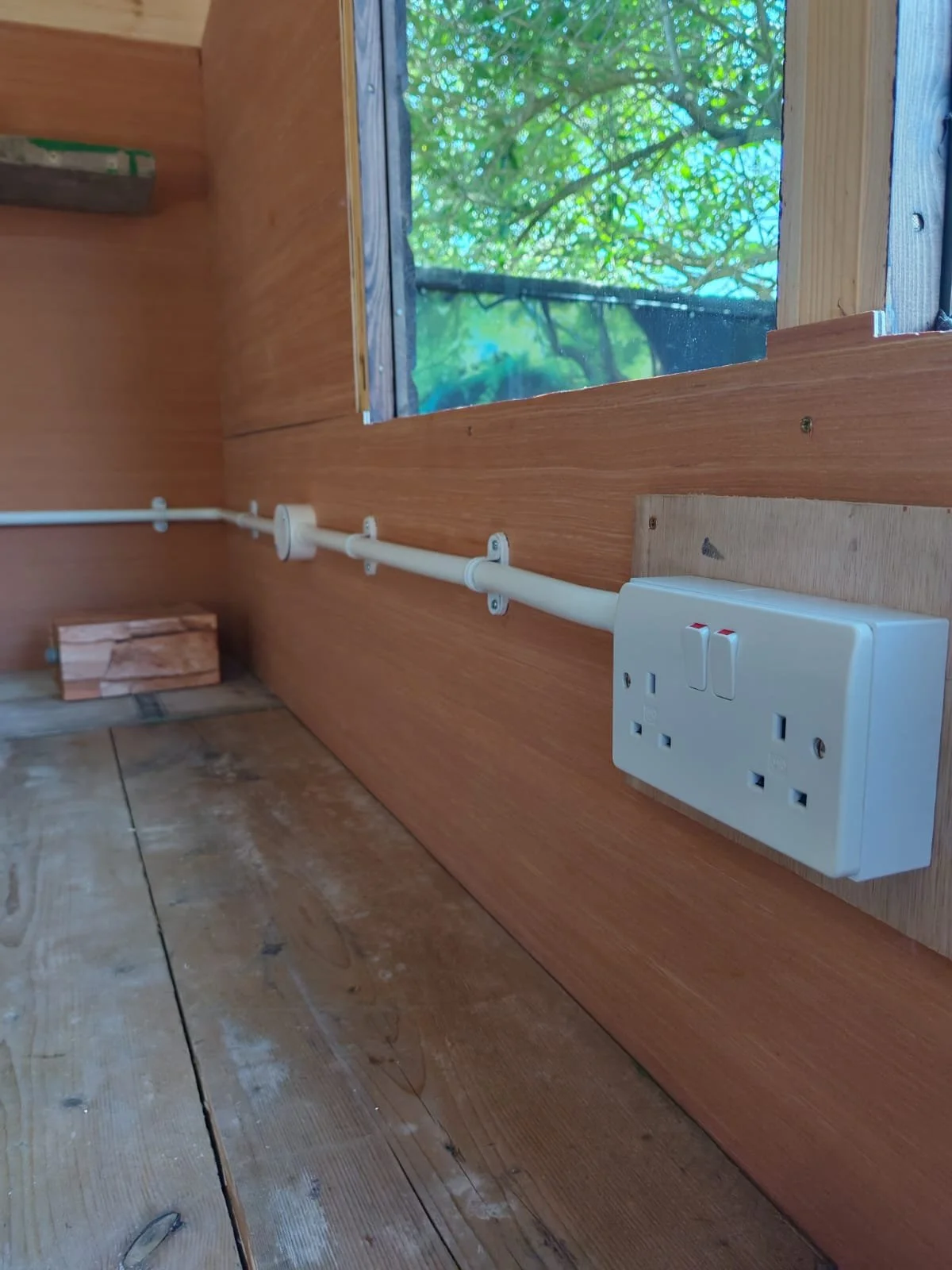 Wooden interior with an electrical outlet, a power switch, a white conduit pipe along the wall, and a window showing green foliage outside.