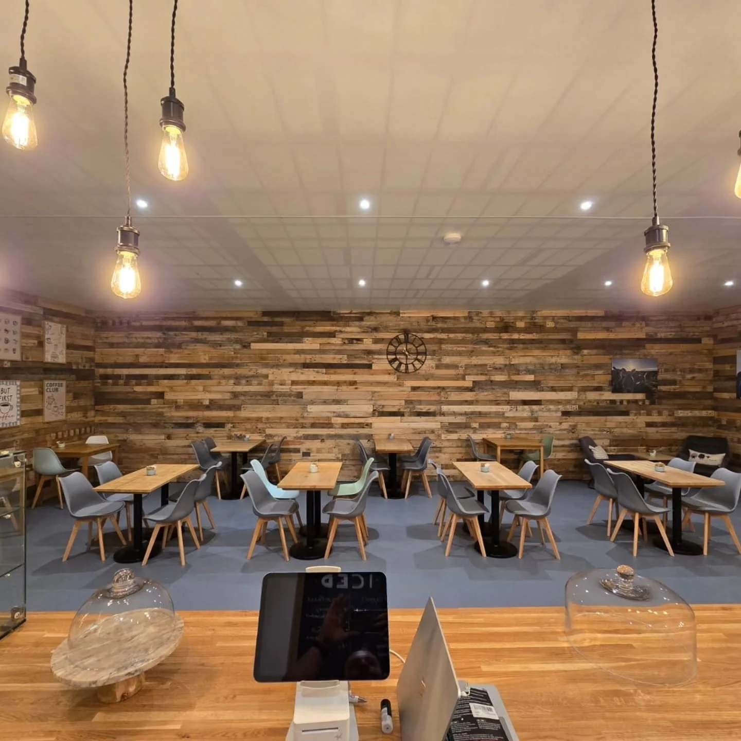 Interior view of a café with wooden wall and tables, gray chairs, pendant lights, and wall art.