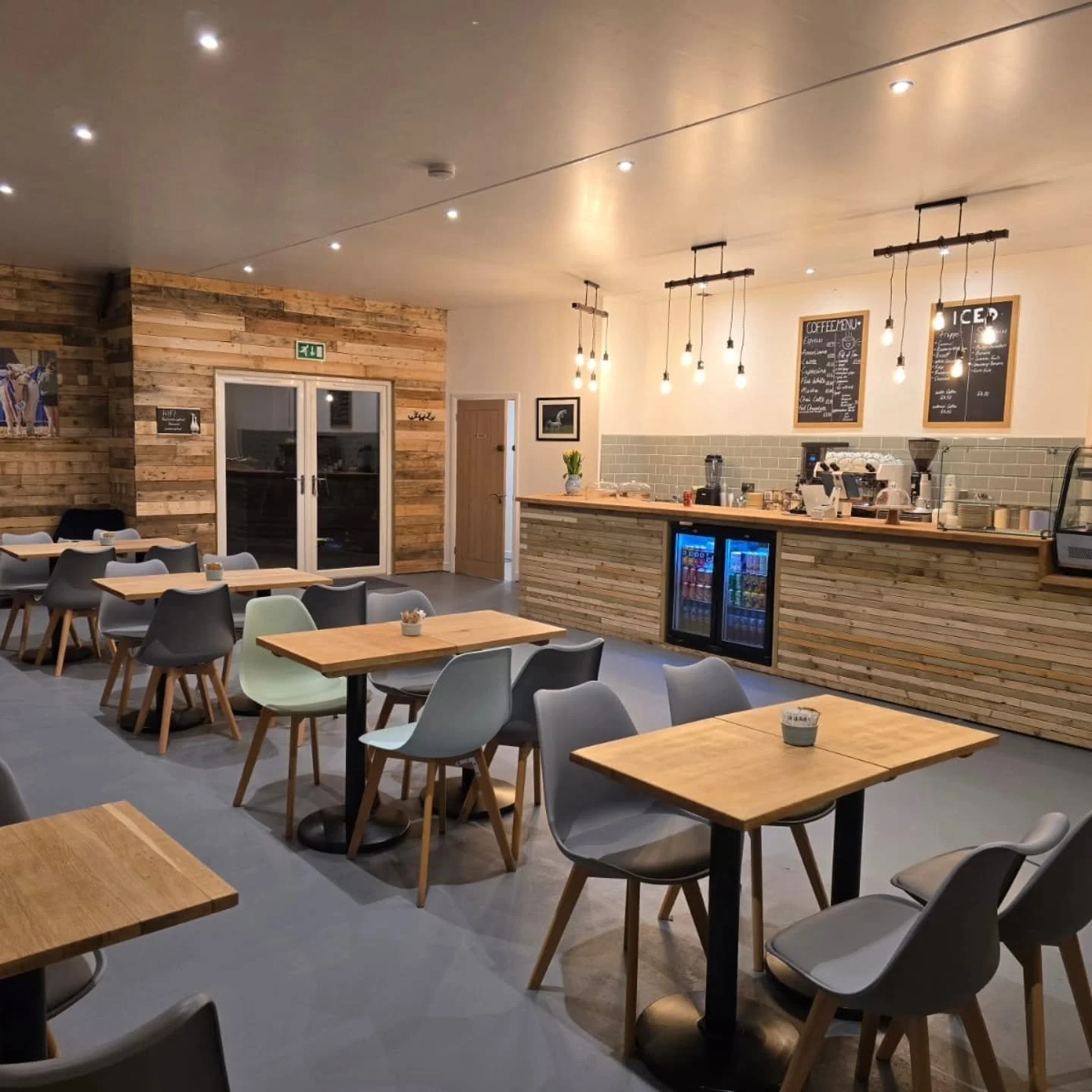 Interior of a cozy coffee shop with wooden tables and chairs, modern lighting, a wooden counter with a variety of coffee equipment, and a beverage refrigerator, with walls decorated in a mix of wood paneling and white paint.