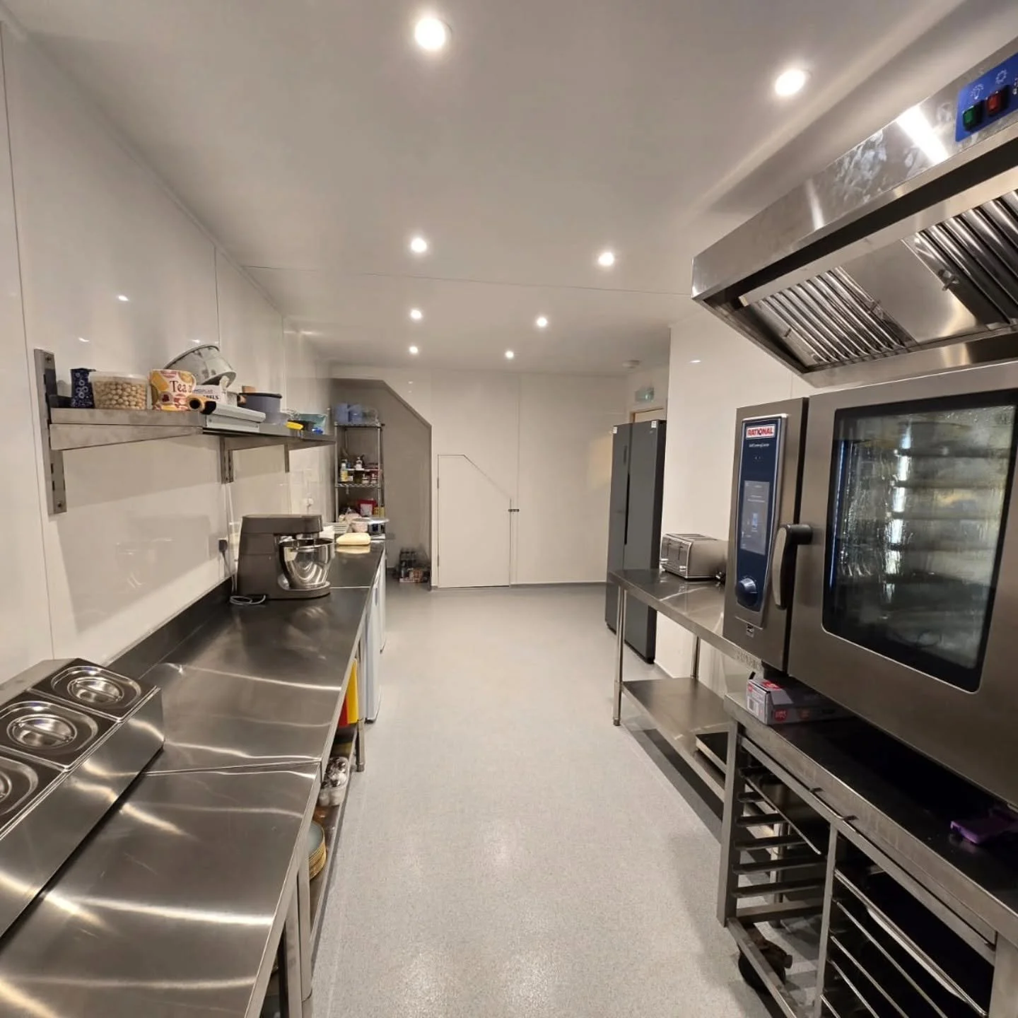A commercial kitchen with stainless steel countertops, appliances, and shelves. Bright lighting and a clean, organized space.