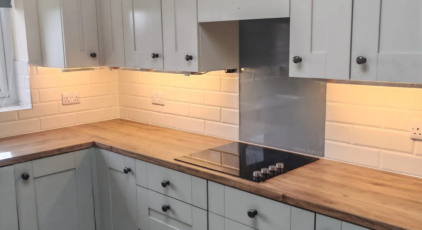 A kitchen with light gray cabinets, a wooden countertop, and a black electric stove. The backsplash features beige subway tiles, and there are multiple electrical outlets on the wall.