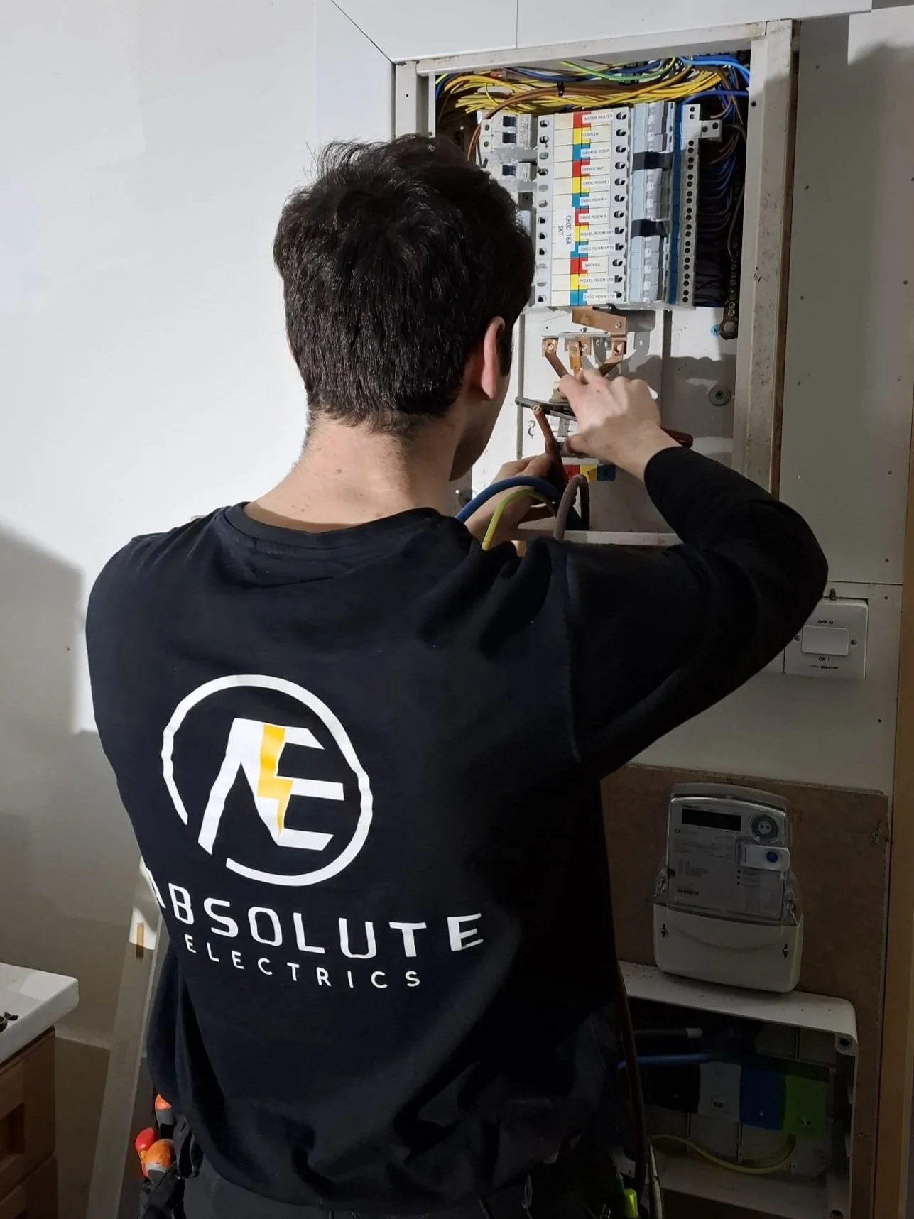 A man working on an electrical panel with various colored wires and switches, wearing a black shirt with white and yellow logo reading 'Absolute Electrics'.