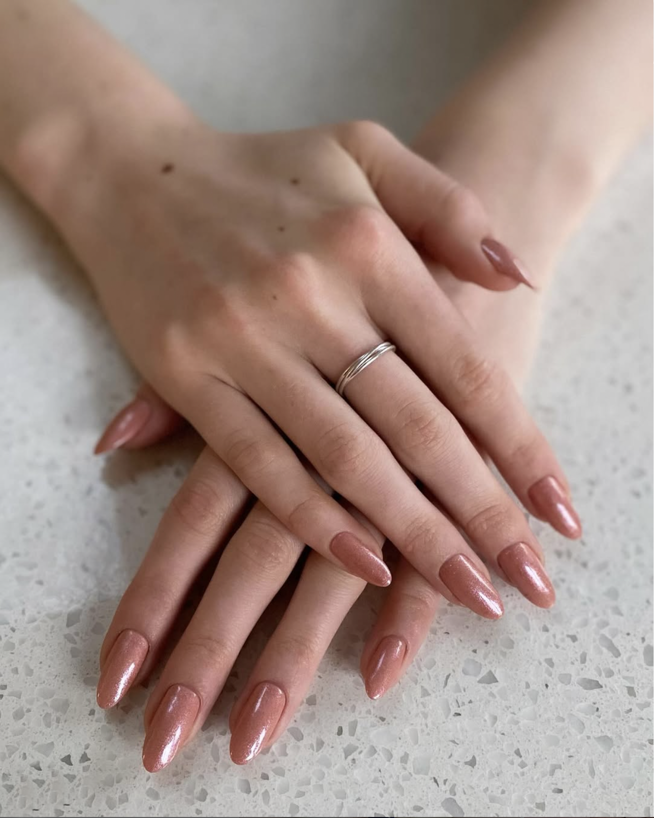 Close-up of two hands with well-manicured, shiny, pink nails. The hand on top has a silver ring on the middle finger. The background is a light-colored surface.