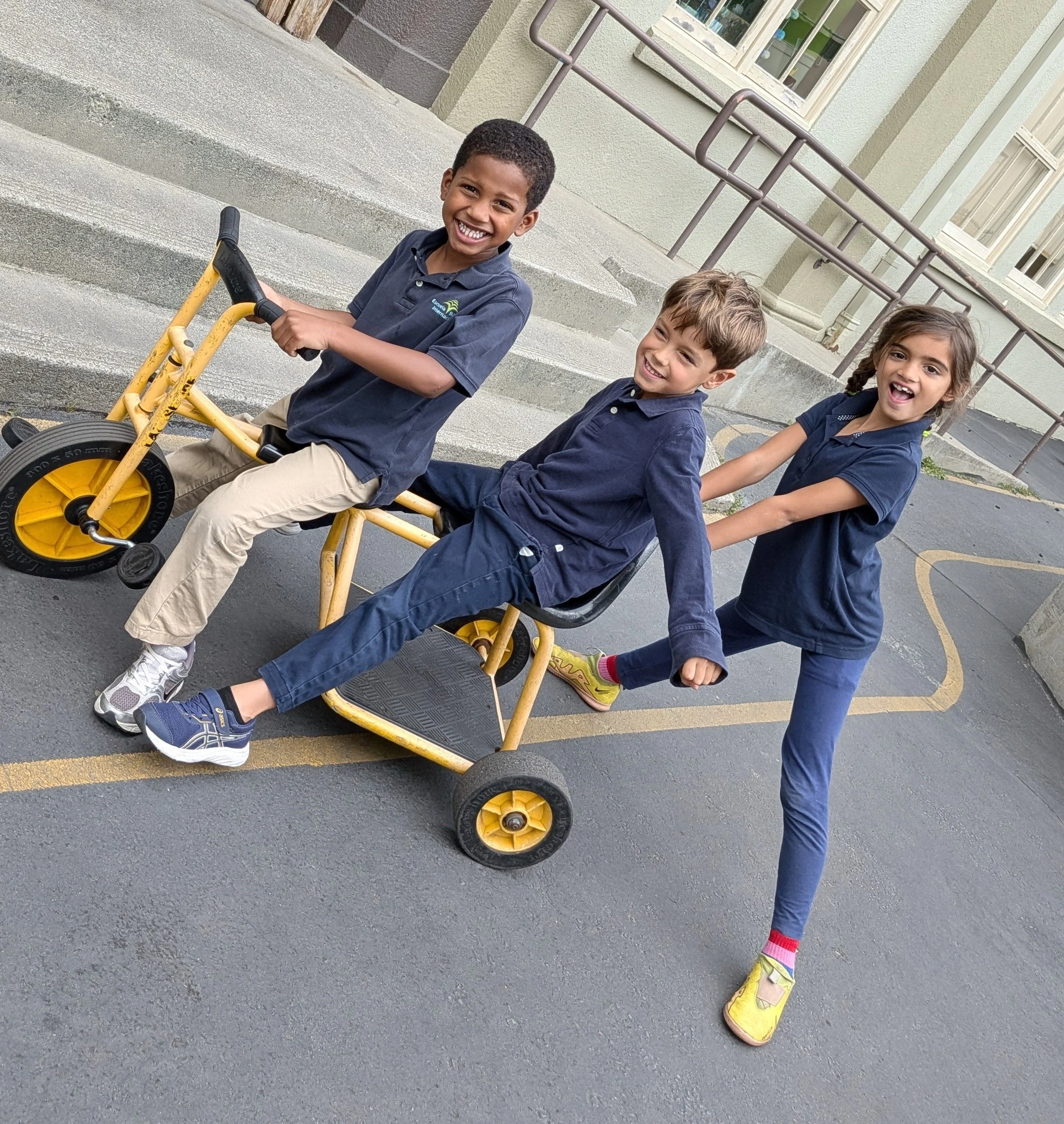 Three children playing outside. One child is sitting on a yellow tricycle, and the other two children are pulling it by holding onto each other's hands, smiling and laughing.