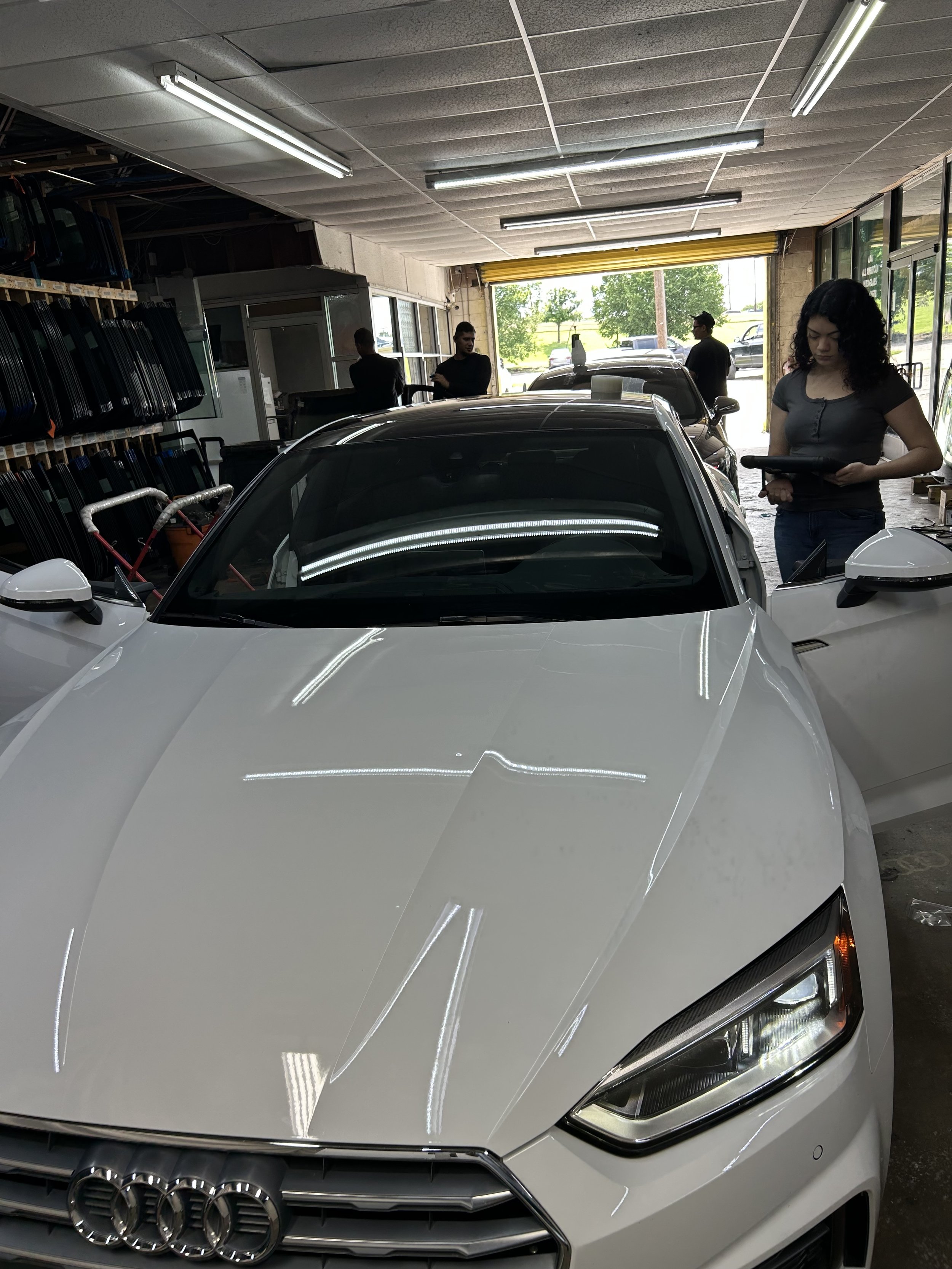 White Audi sports car inside an auto shop with people working on cars and shelves of car parts in the background.
