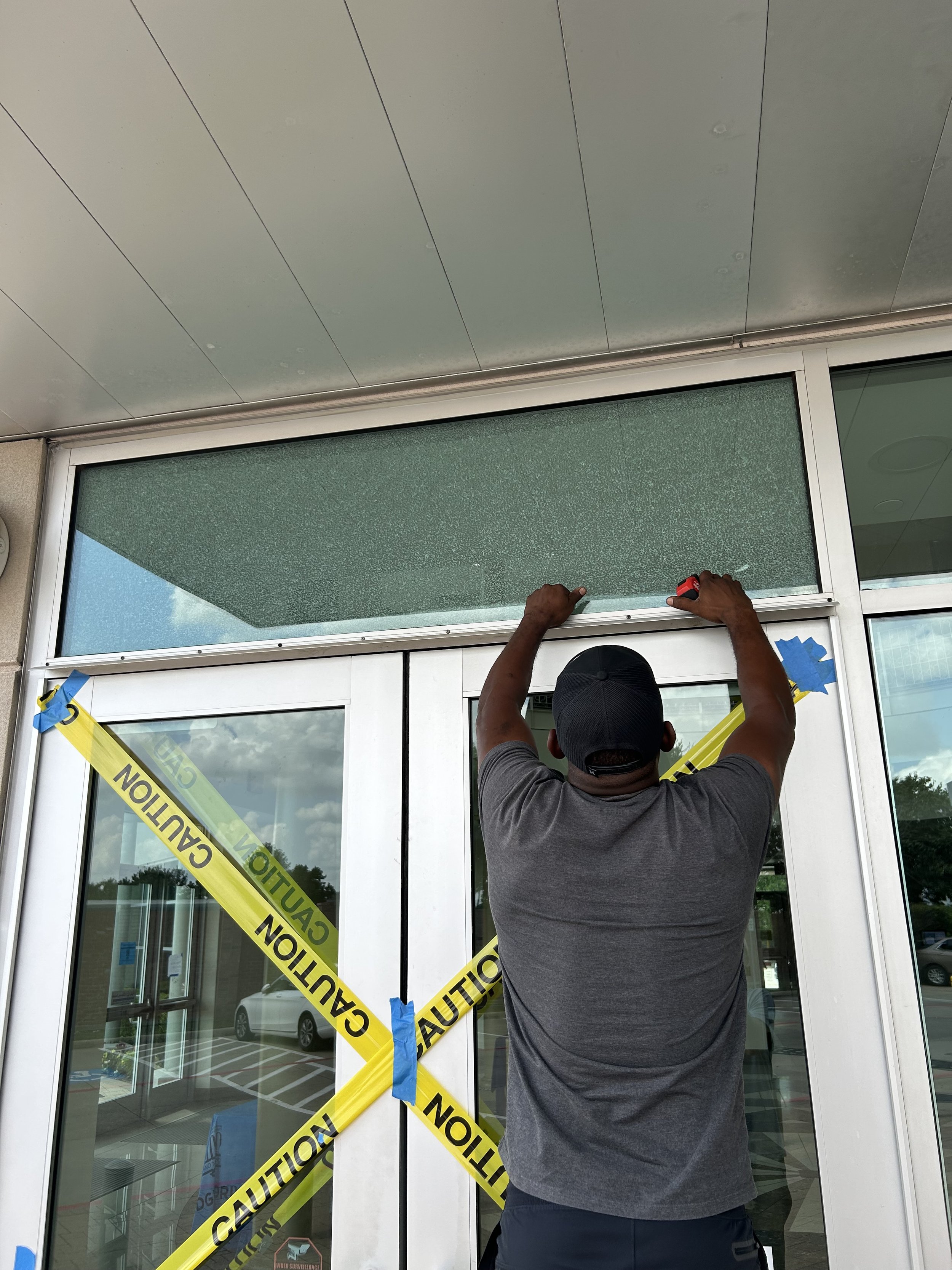 A man installing or repairing a glass panel above glass doors at a building entrance, with caution tape across the doors.