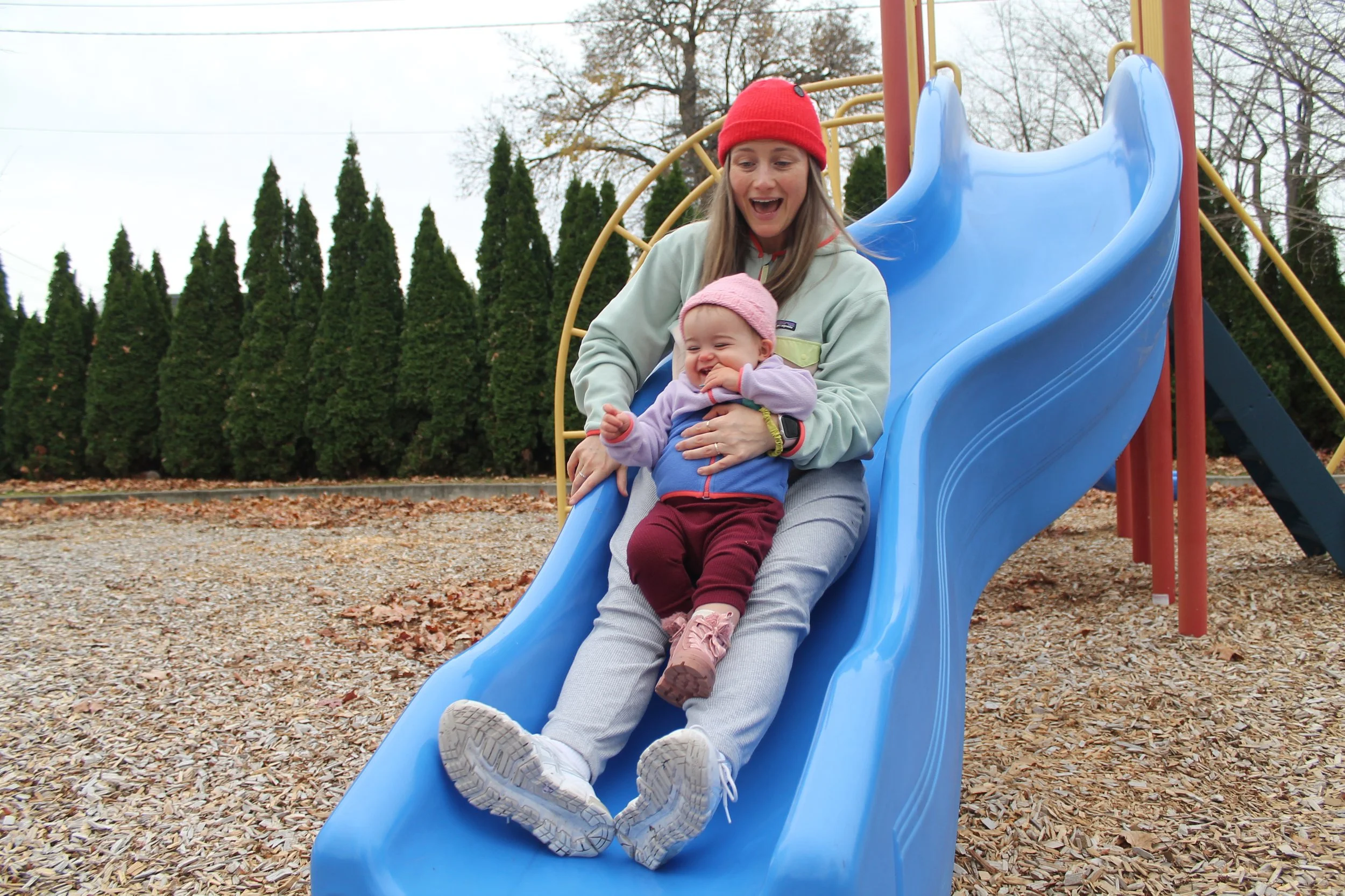 A woman and a young girl sitting at the top of a blue slide at a playground, smiling and enjoying fun time together.
