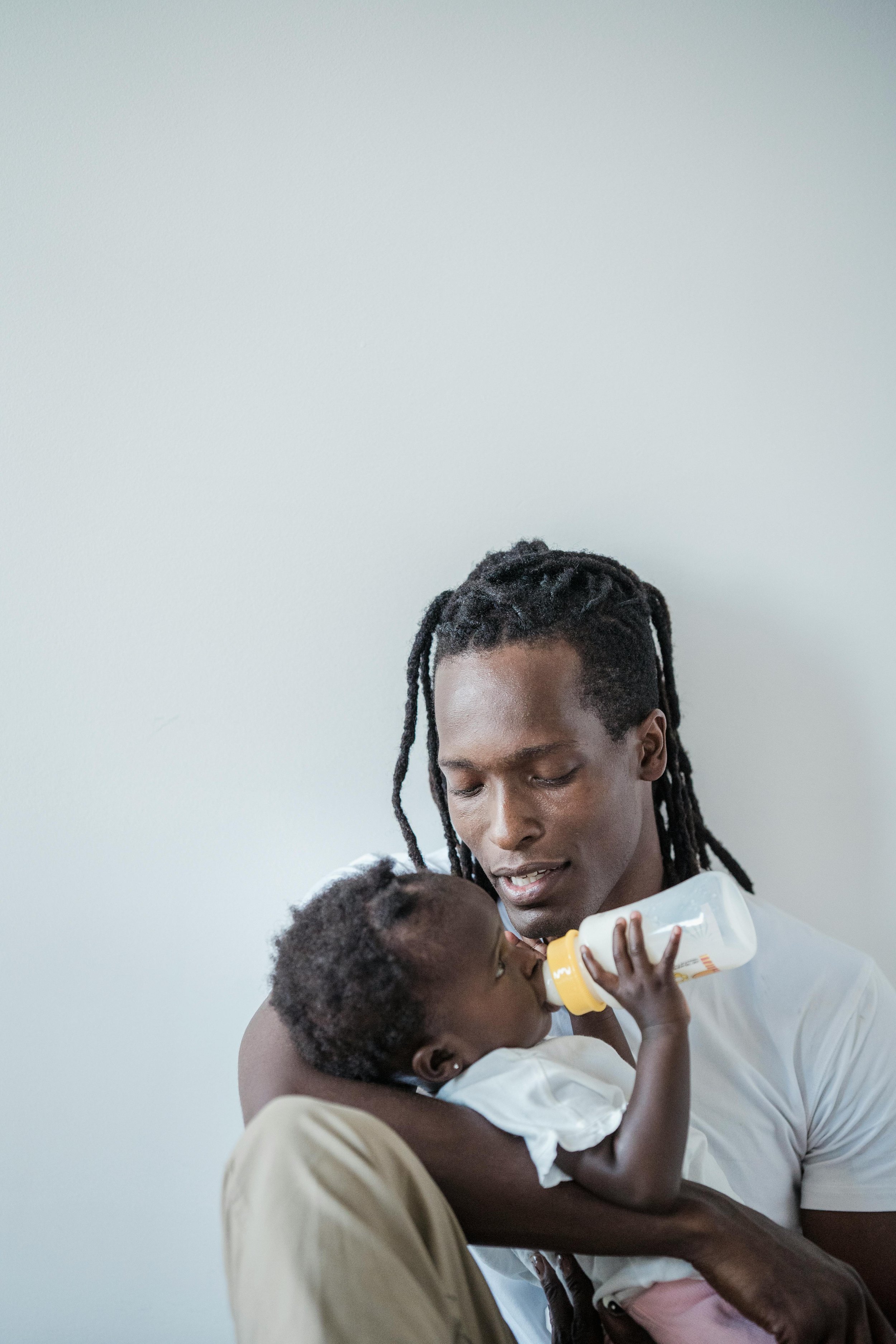 A man with dreadlocks feeds a young child with a baby bottle, holding the child close to his chest, against a plain white background.