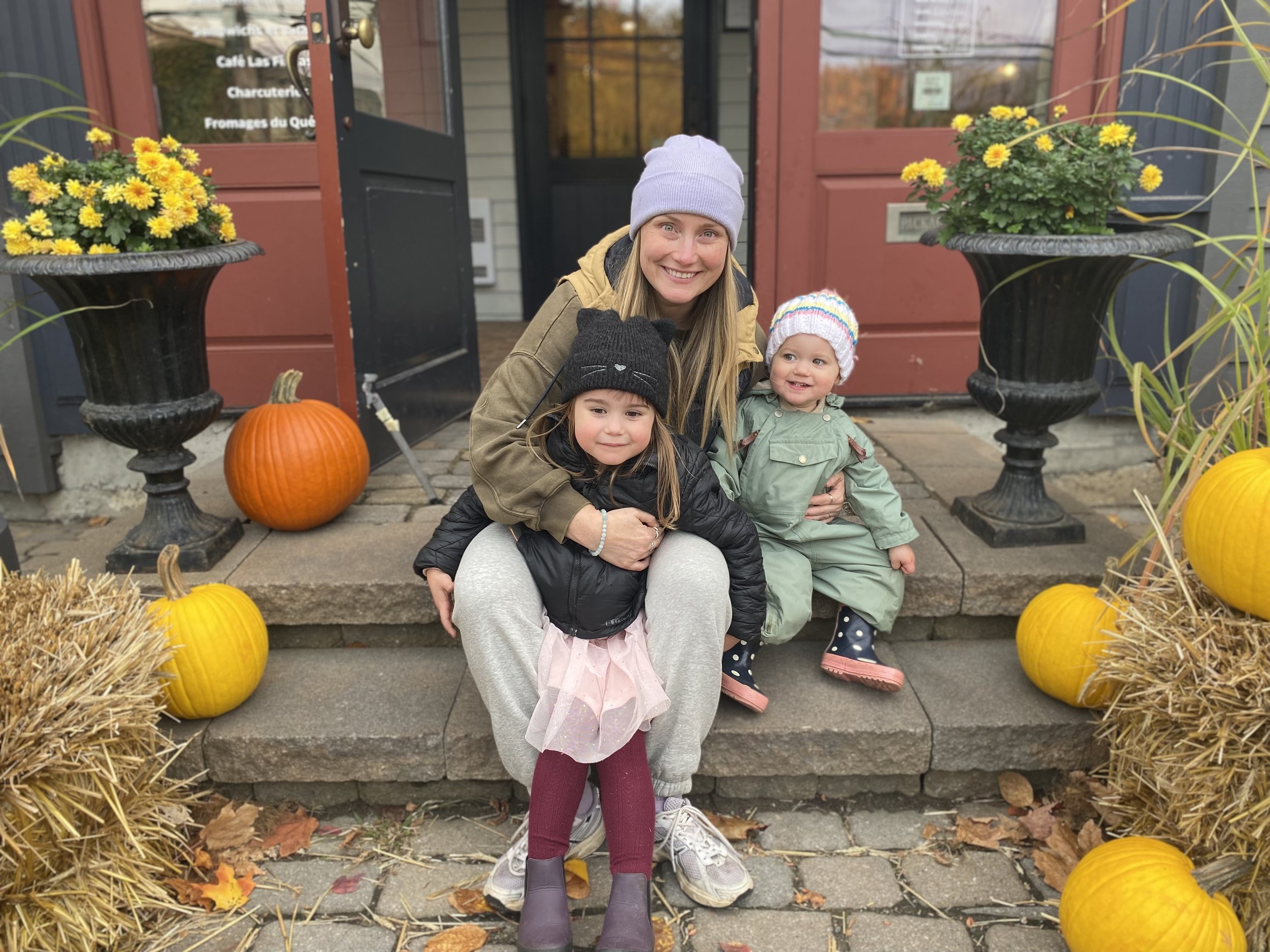 A woman smiling with two young girls sitting on front steps outside a building decorated with pumpkins and hay bales for fall.