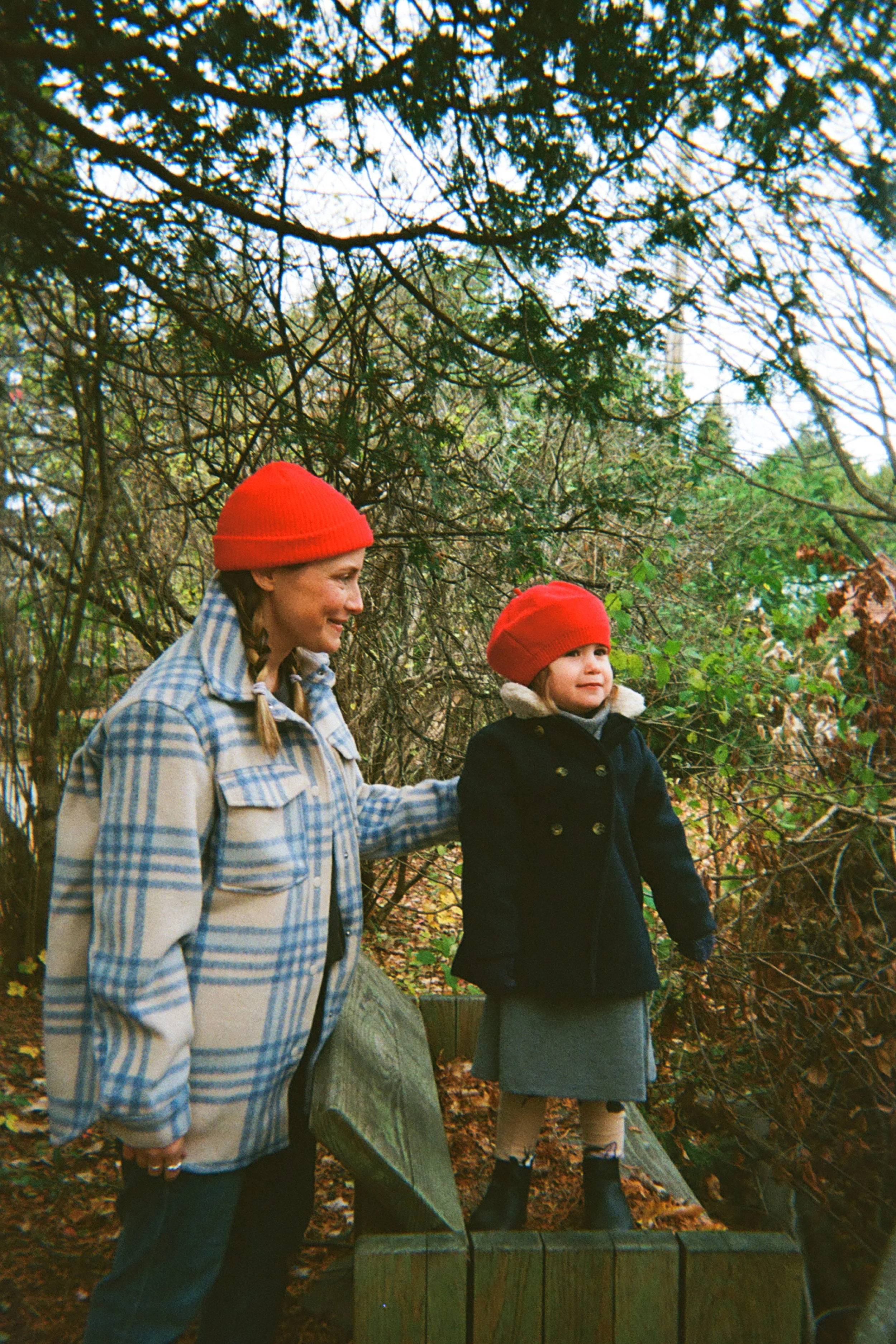 A woman and a young girl standing on a wooden platform outdoors in a wooded area, both wearing red hats, with the woman smiling at the girl who is looking into the distance.