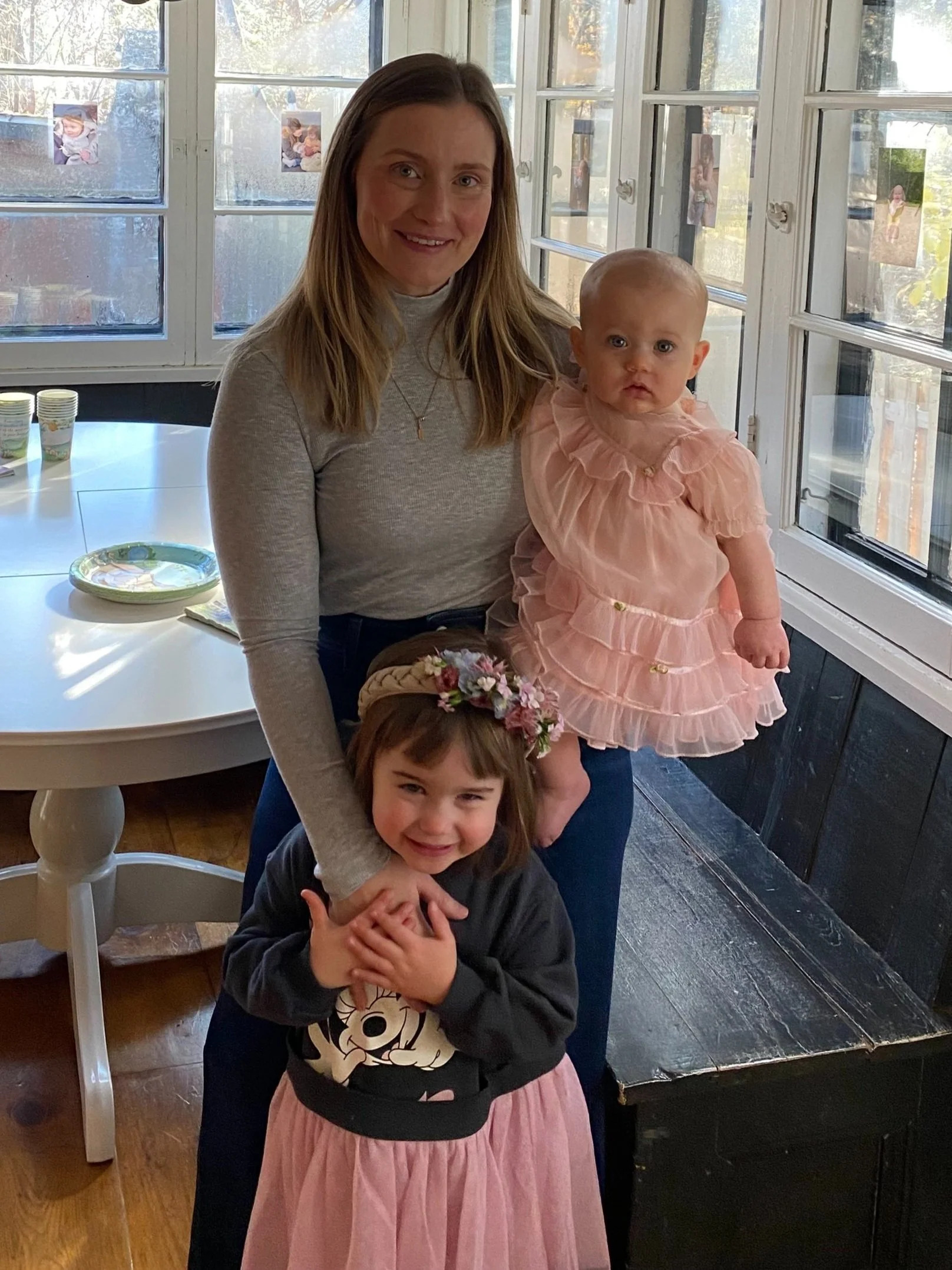 A woman with two young girls inside a house with large windows and photos on the window panes. The woman is smiling and has straight brown hair, wearing a gray long-sleeve shirt.