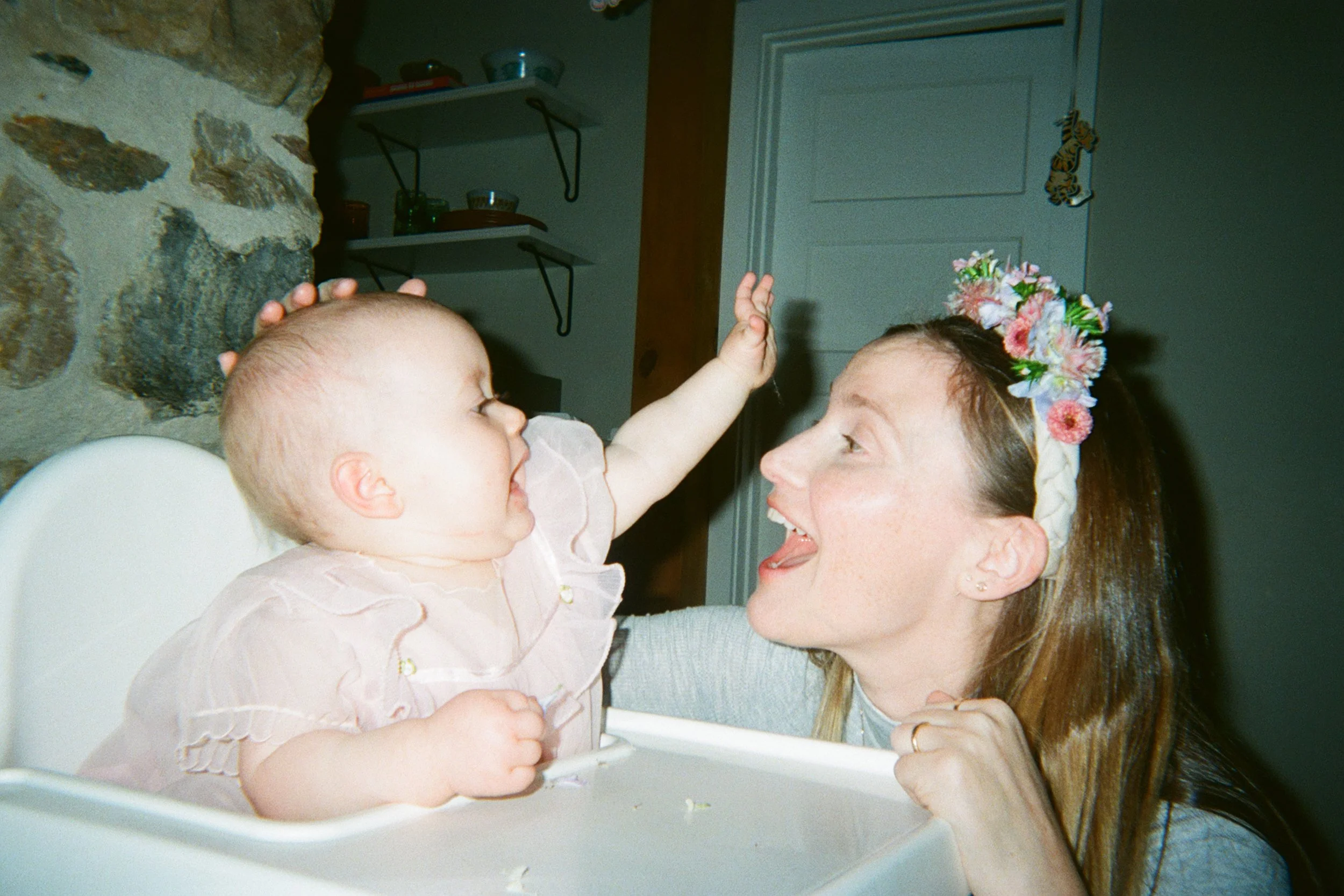 A woman with a floral headband smiling and reaching out to a baby sitting in a high chair, touching the woman's forehead with her hand.