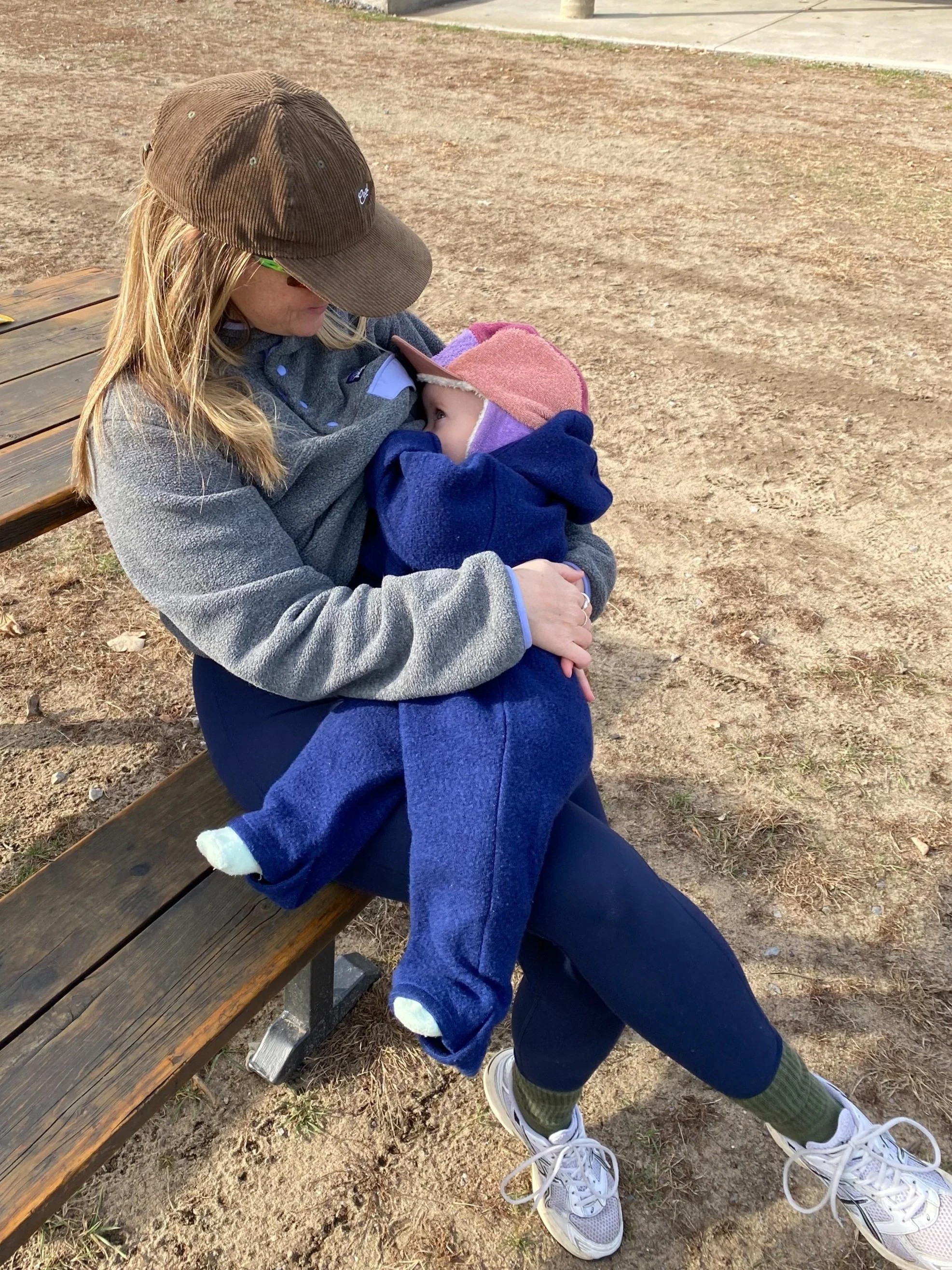 A woman is sitting on a wooden park bench holding a young child on her lap. They are hugging each other, with the woman looking down at the child.