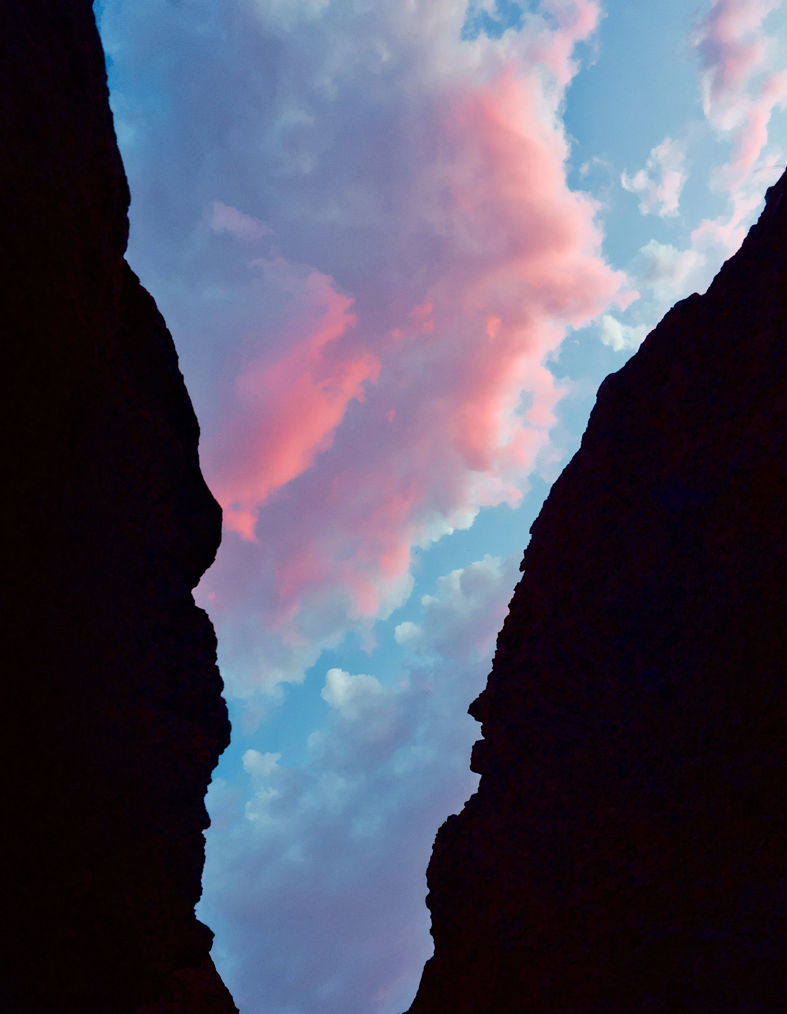 A view of the sky with pink and blue clouds seen through a deep canyon or gorge with dark rock walls on either side.