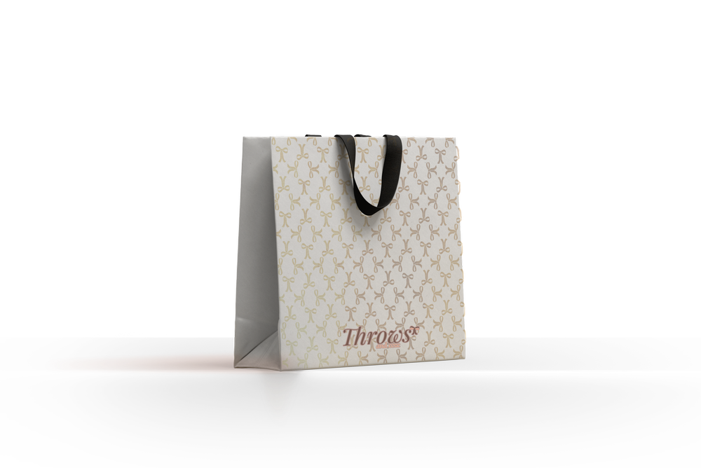 A cream Throws & Coze shopping bag with a monogram pattern and black ribbon handles.