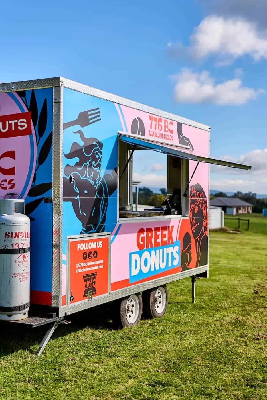 A colorful food truck painted with Greek mythology themes, advertising Greek donuts, parked on grass under a partly cloudy sky.